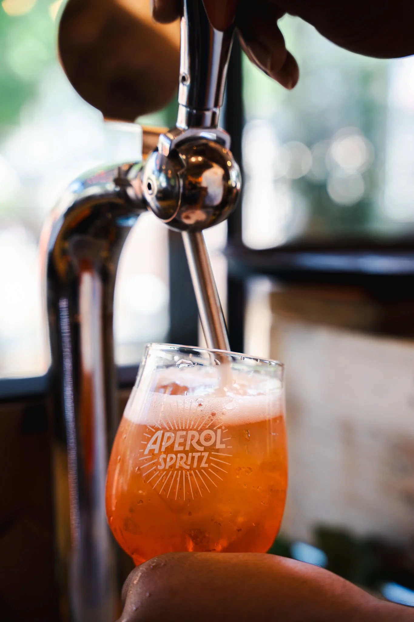 A glass of Aperol Spritz being filled with orange-colored liquid from a tap, with the glass labeled 'Aperol Spritz'. Bright and blurred outdoor background.