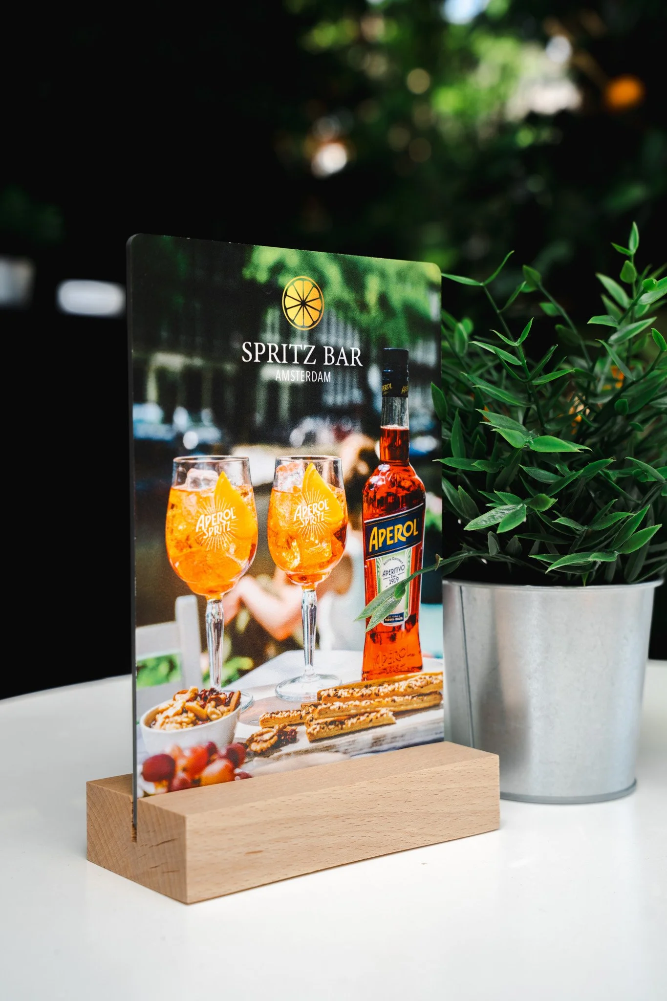 A table sign for Spritz Bar Amsterdam featuring an image of two Aperol Spritz drinks, a bottle of Aperol, and some snacks, with a potted plant beside it.