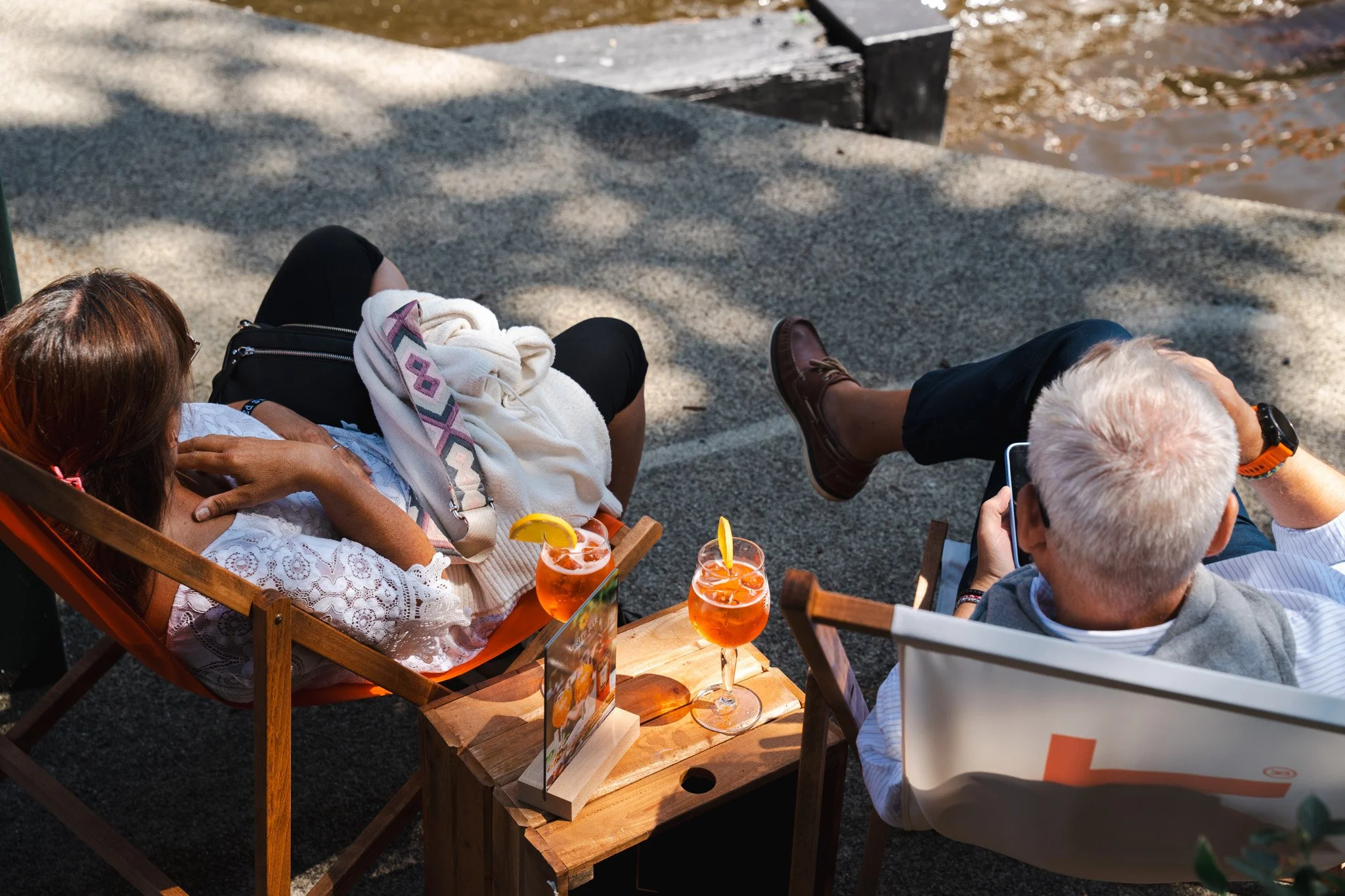 Two people relaxing on a riverside dock, one woman lying on a lounge chair with a blanket and two drinks on a small wooden table, and one man sitting in a high-back chair looking at his phone, with water visible in the background.