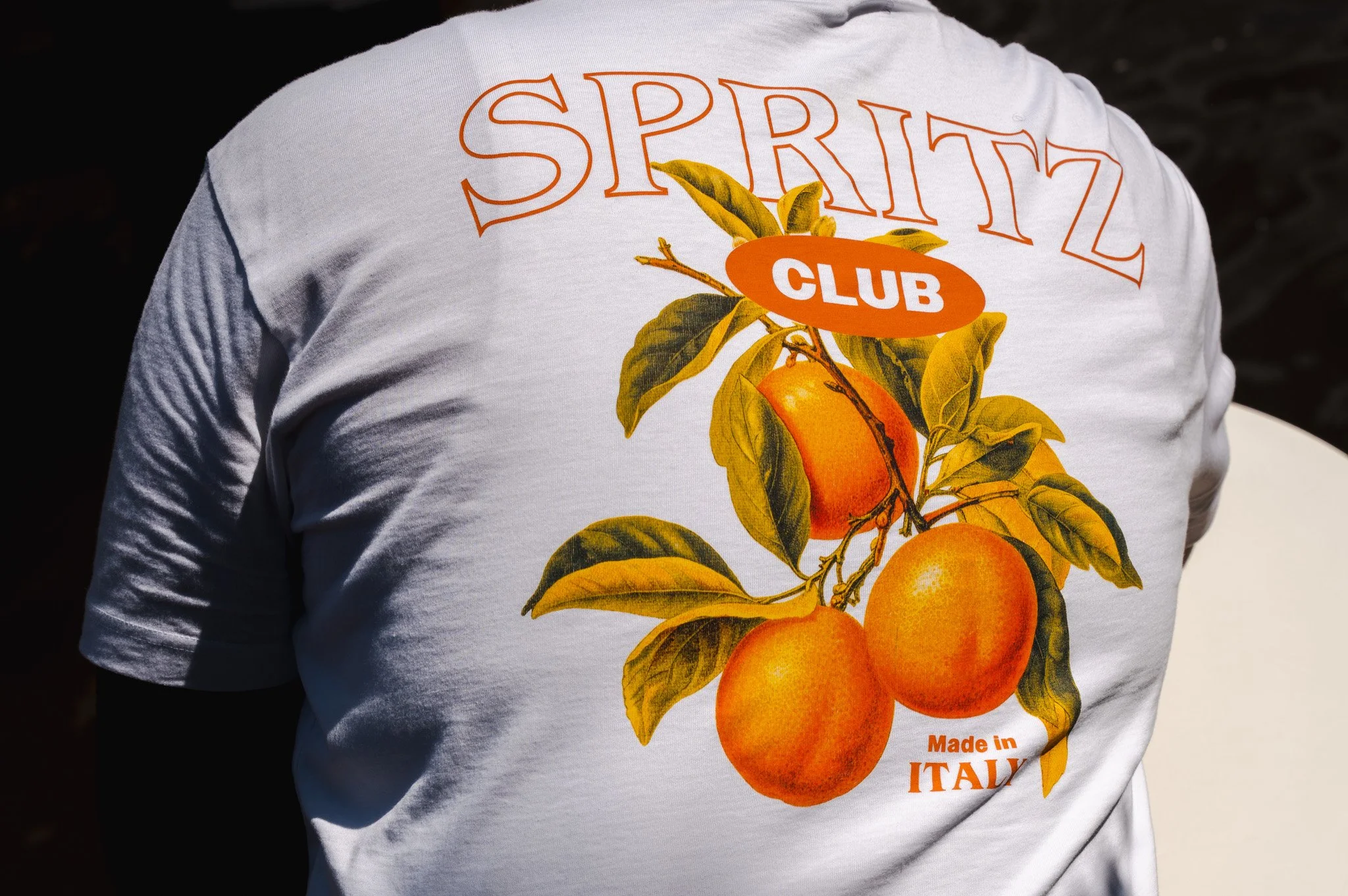 Person wearing a white T-shirt with a graphic of orange fruit and leaves, the text 'SPRITZ CLUB' and 'Made in Italy' printed on the back.