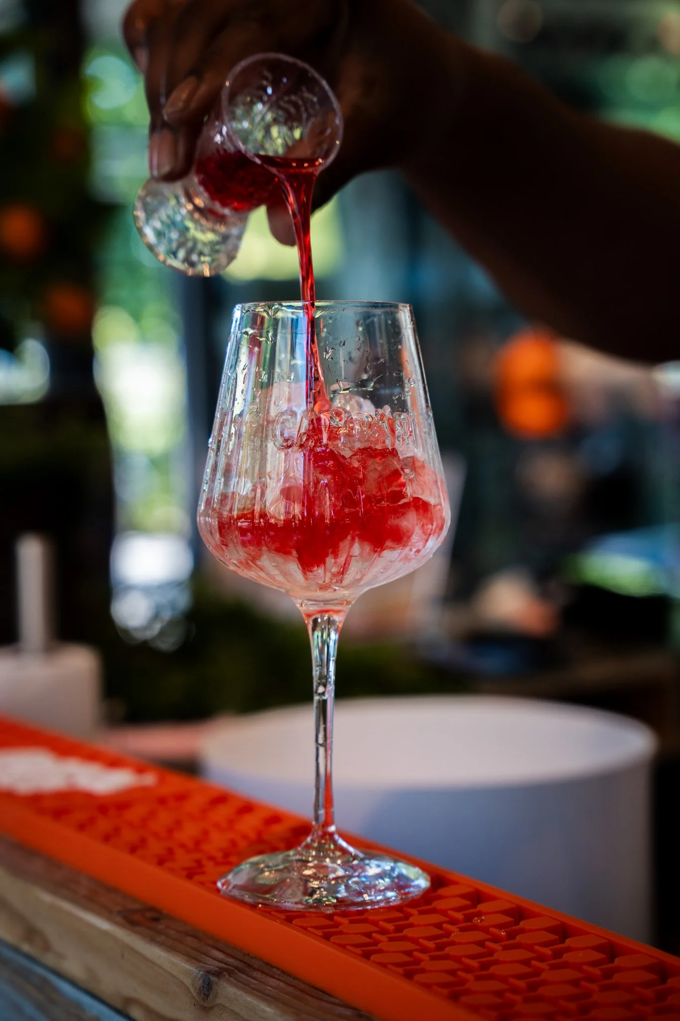 A hand pouring red liquid into a tall glass with ice cubes, on an orange bar mat.