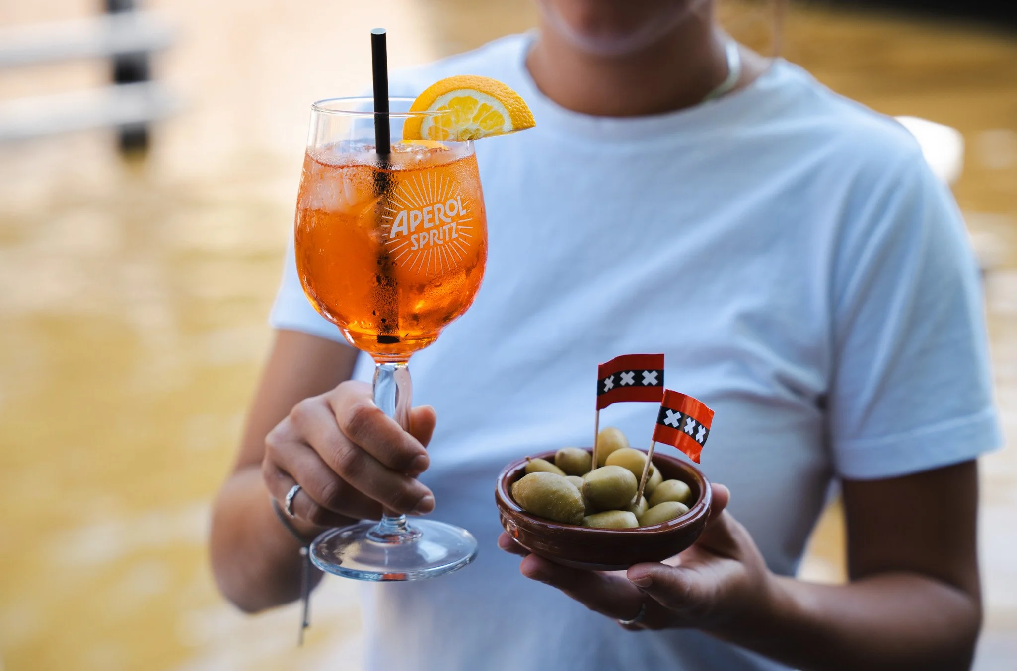 Person holding a glass of Aperol Spritz with lemon wedge and a small bowl of olives with flags, outdoors near water