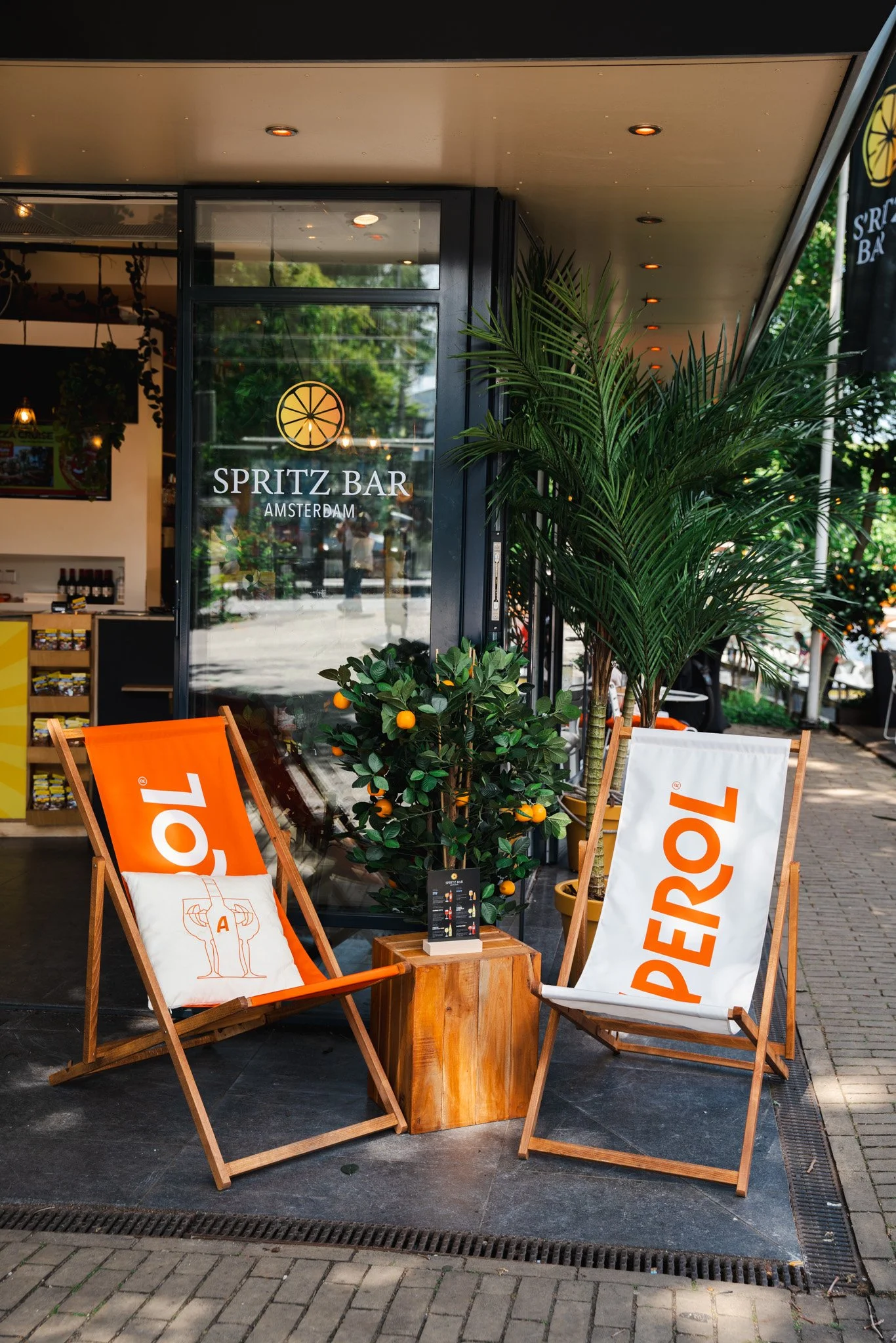 Outdoor seating area outside Spritz Bar, Amsterdam with two wooden chairs with orange and white fabric, a small wooden table, potted orange tree, tall plant, and the bar's glass door entrance.