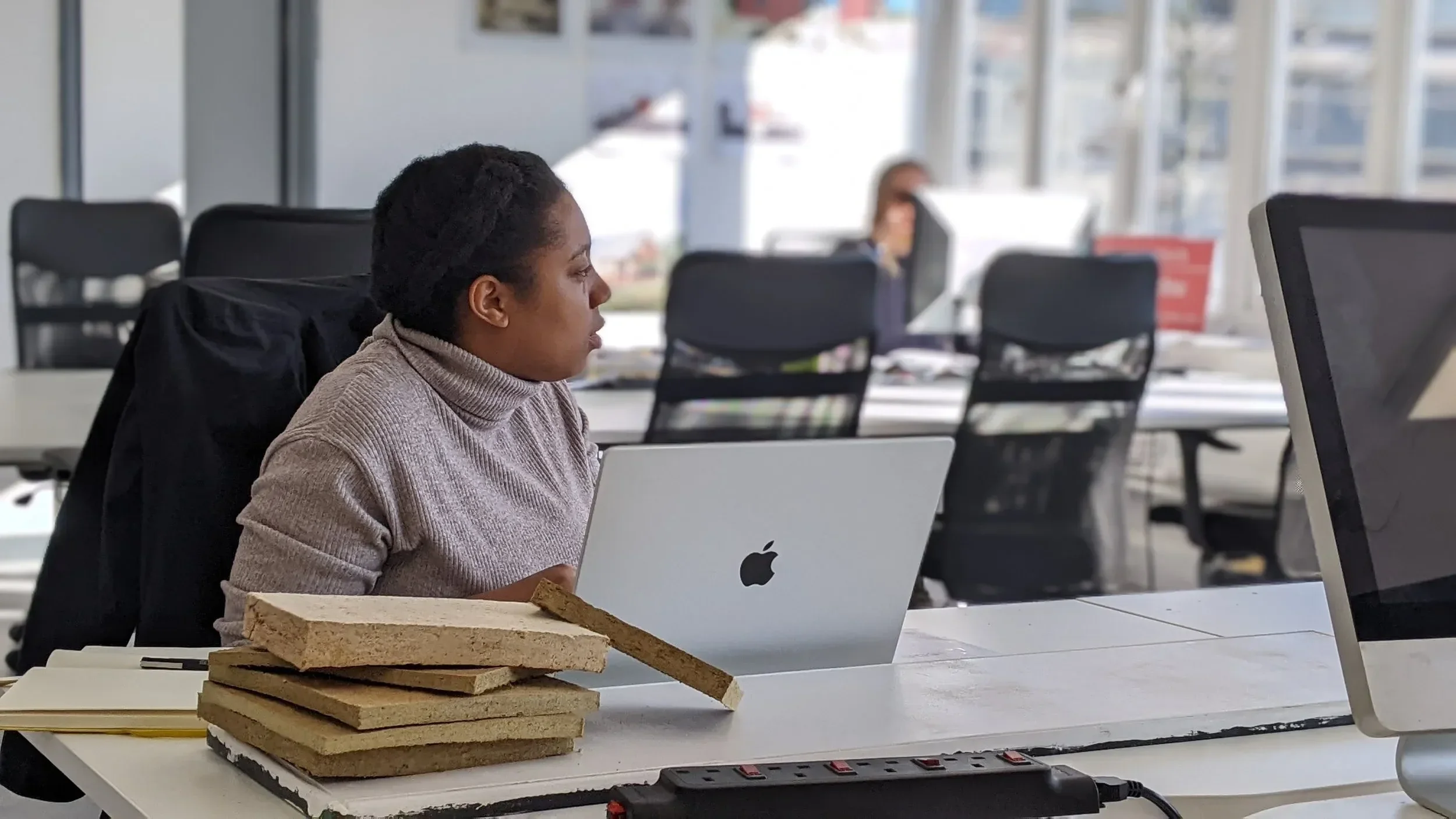 A woman working at a desk with an Apple laptop and a computer monitor in an office with large windows. There are stacks of tiles and a notebook on the desk, and other people working at desks in the background.