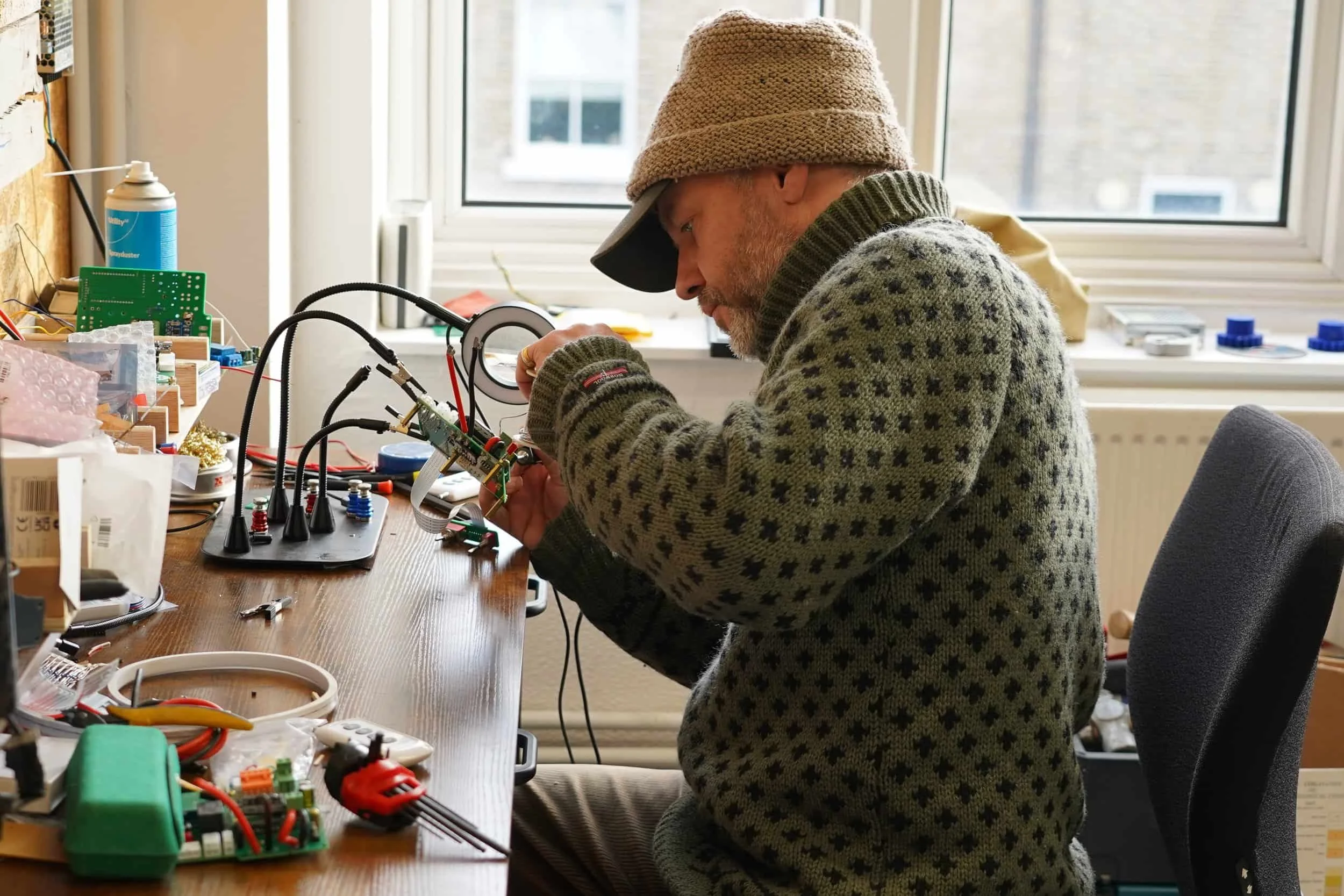 Man working on electronic circuit board in a workshop, seated at a cluttered wooden table with various tools and components, near a window.