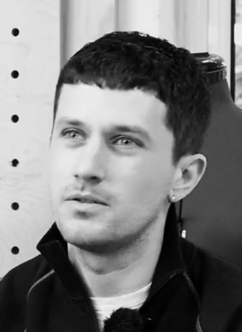 Black and white photo of a young man with short dark hair, wearing a dark hoodie and a small earring on his left ear, looking directly at the camera.
