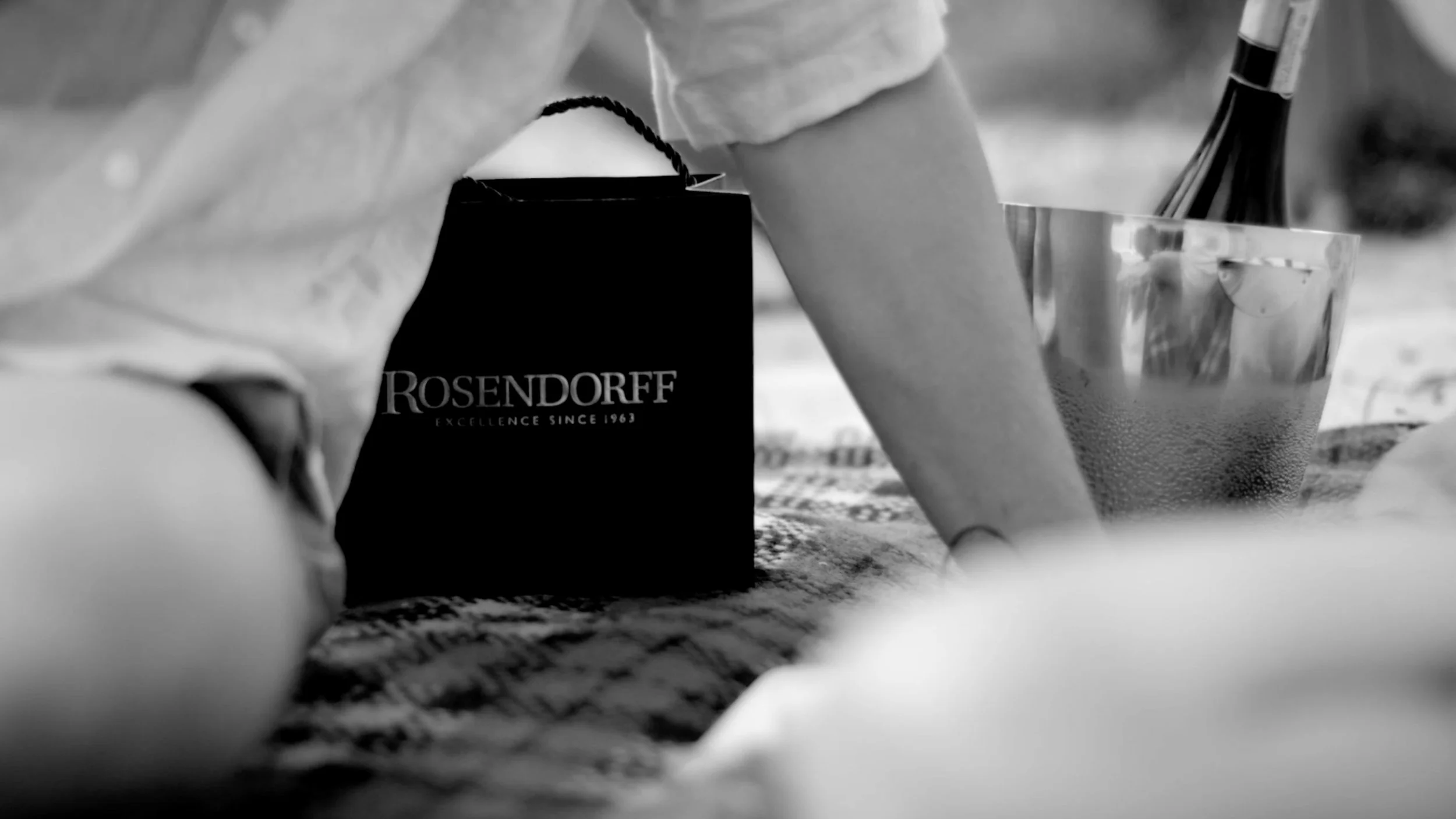 A person's arm and hand are visible, holding a black paper bag with the brand name 'Rosendorff' on it, placed on a patterned fabric surface. Next to the bag is a metal ice bucket with a bottle of wine inside.
