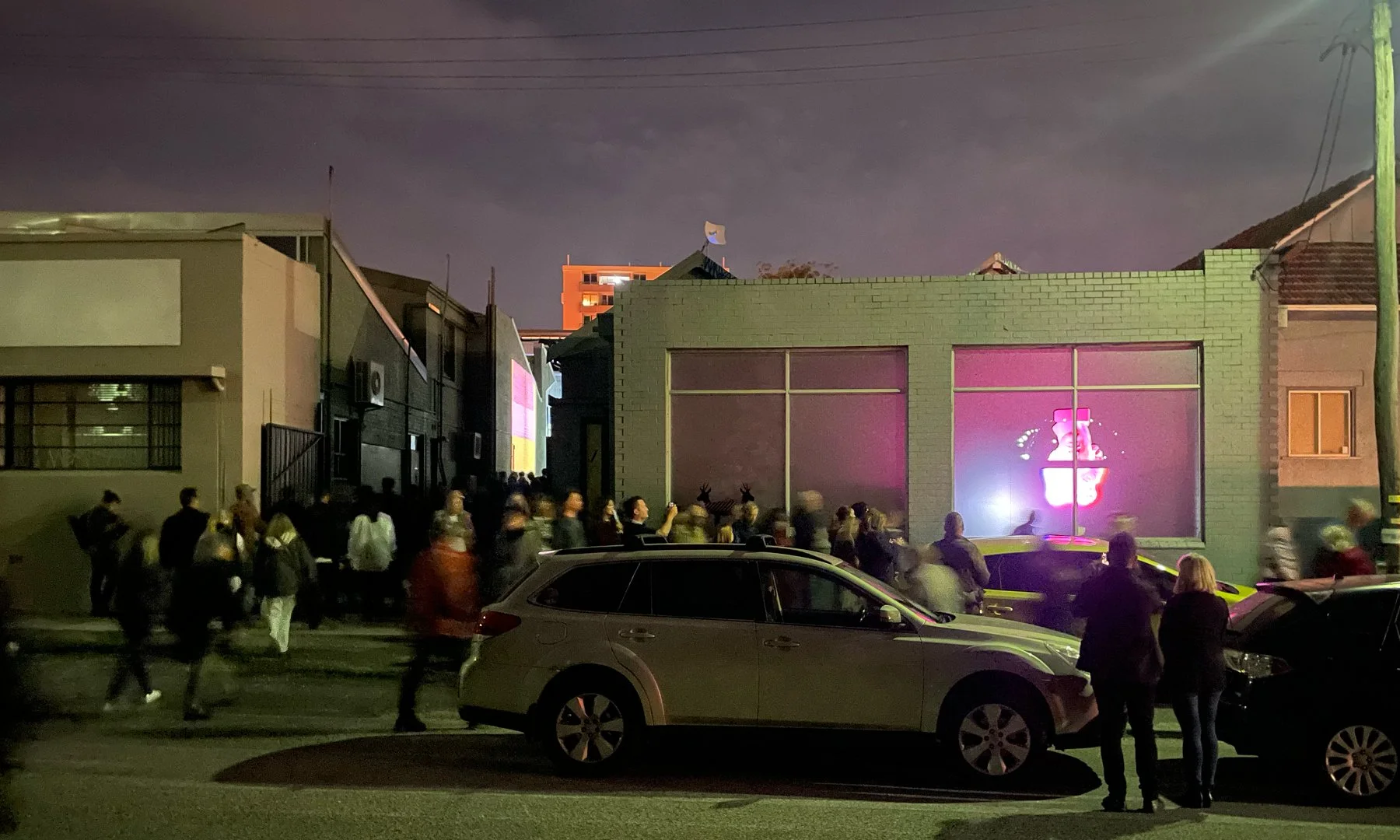 Pickle District - Night scene with a crowd of people outside a building with neon lights, parked cars, and a purple-lit decoration on the window.