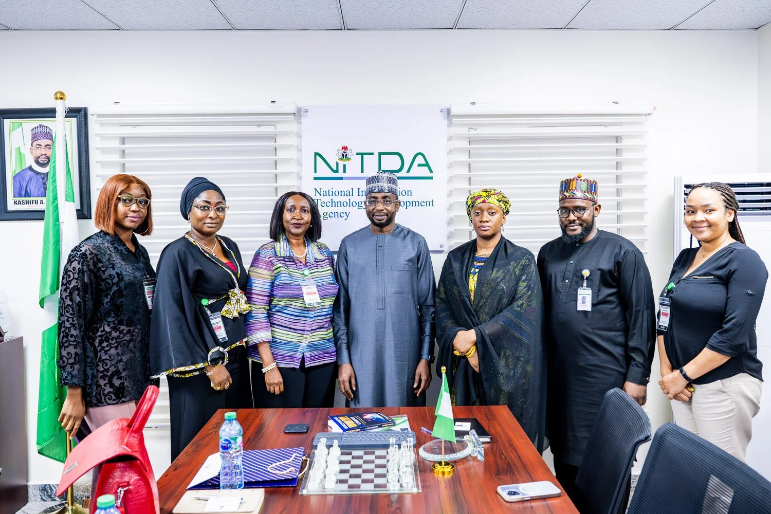 A group of seven people standing in a conference room in front of a sign that reads 'NITDA' for the National Information Technology Development Agency. The group includes six women and one man, all dressed in formal or traditional attire. There is a table in front of them with a chessboard, bottled water, a phone, notebooks, and a small Nigerian flag.