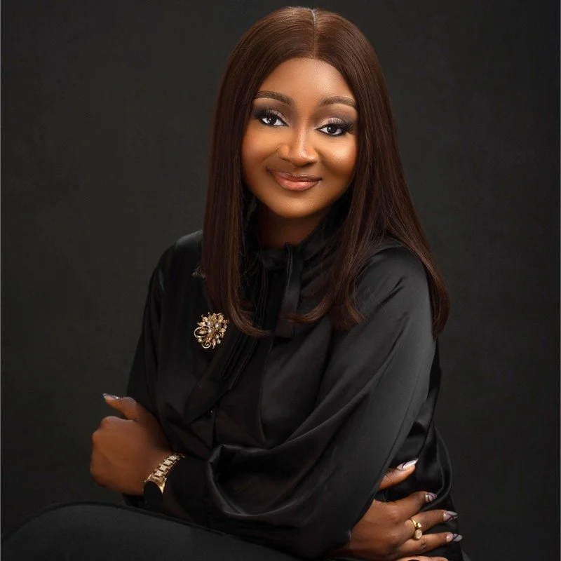 Portrait of a woman with straight brown hair and light makeup, wearing a black satin blouse with a brooch and gold jewelry, smiling with arms crossed against a dark background.