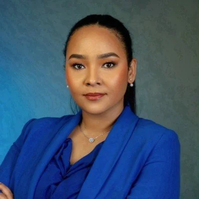 A woman with dark hair pulled back, wearing a blue blazer and a matching blouse, standing against a gradient gray background.