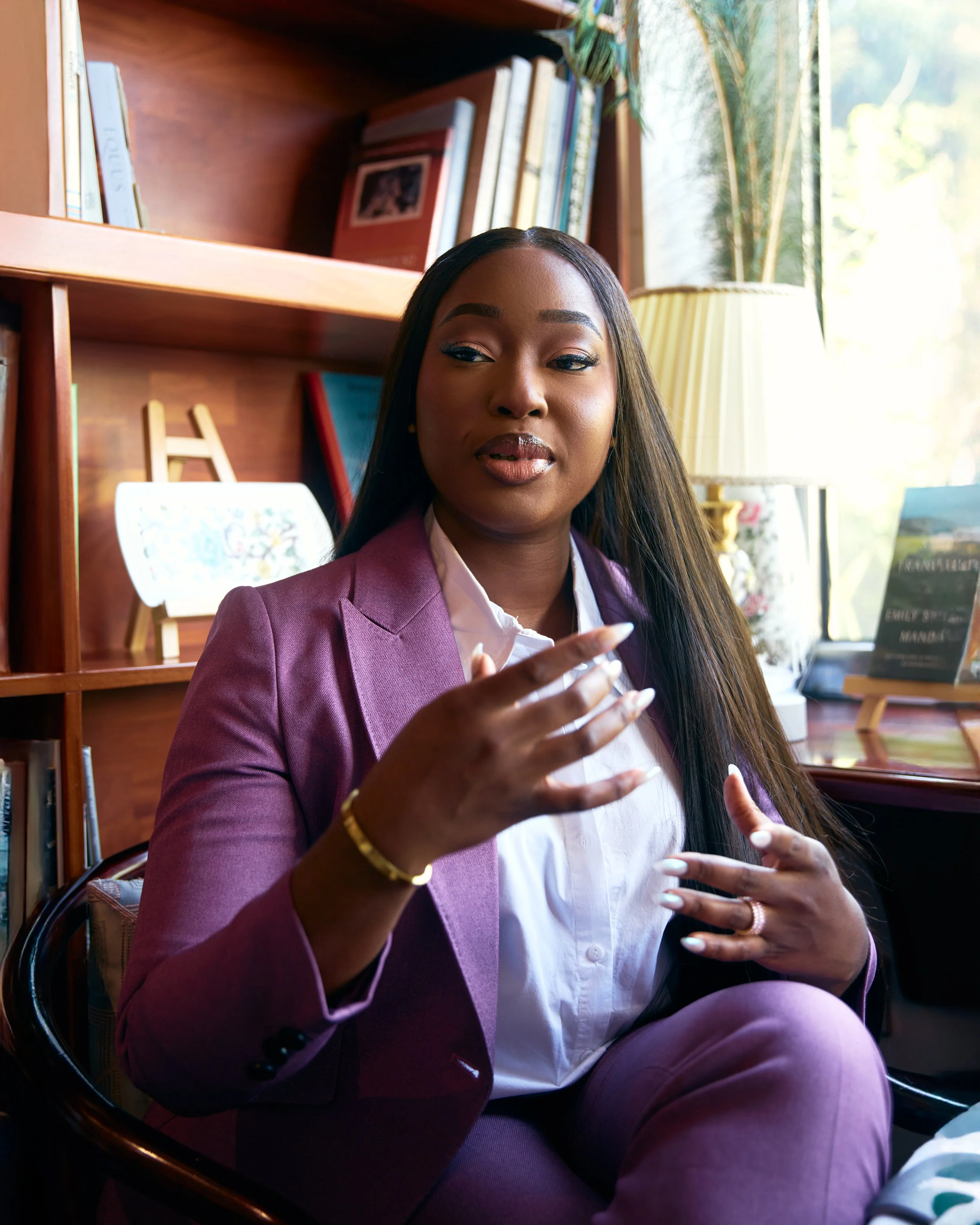 A woman in a purple blazer and pants sitting in a cozy room with a wooden bookshelf, window, and lamp behind her, engaged in conversation or presentation.