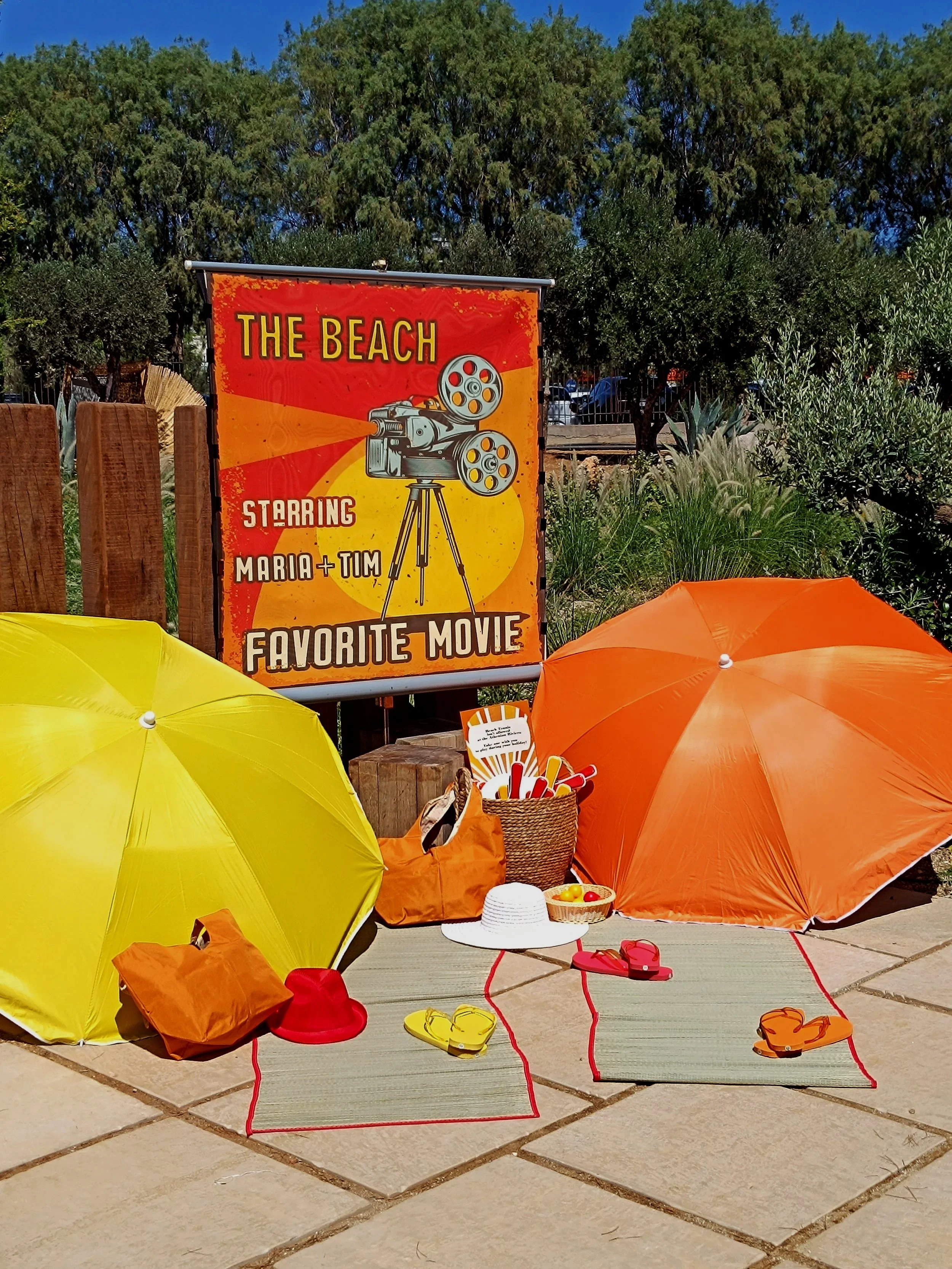 A summer beach scene with yellow and orange umbrellas, beach mats, hats, sandals, a basket of snacks, and a sign that reads 'The Beach Starring Maria + Tim Favorite Movie' with an illustration of a vintage movie camera.