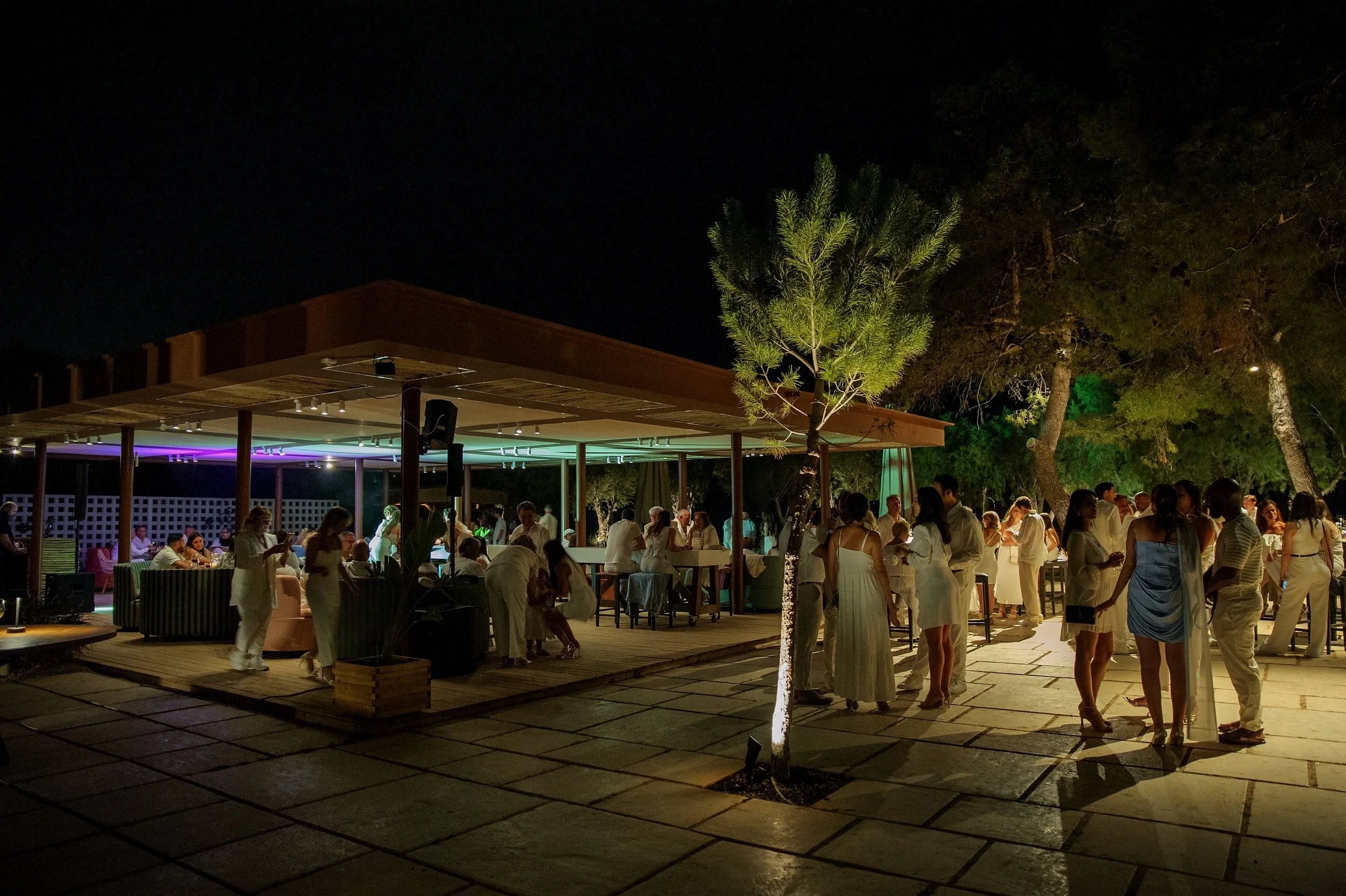 Nighttime outdoor party with people socializing, illuminated by ambient lighting, under a covered patio area and around trees on a tiled patio.