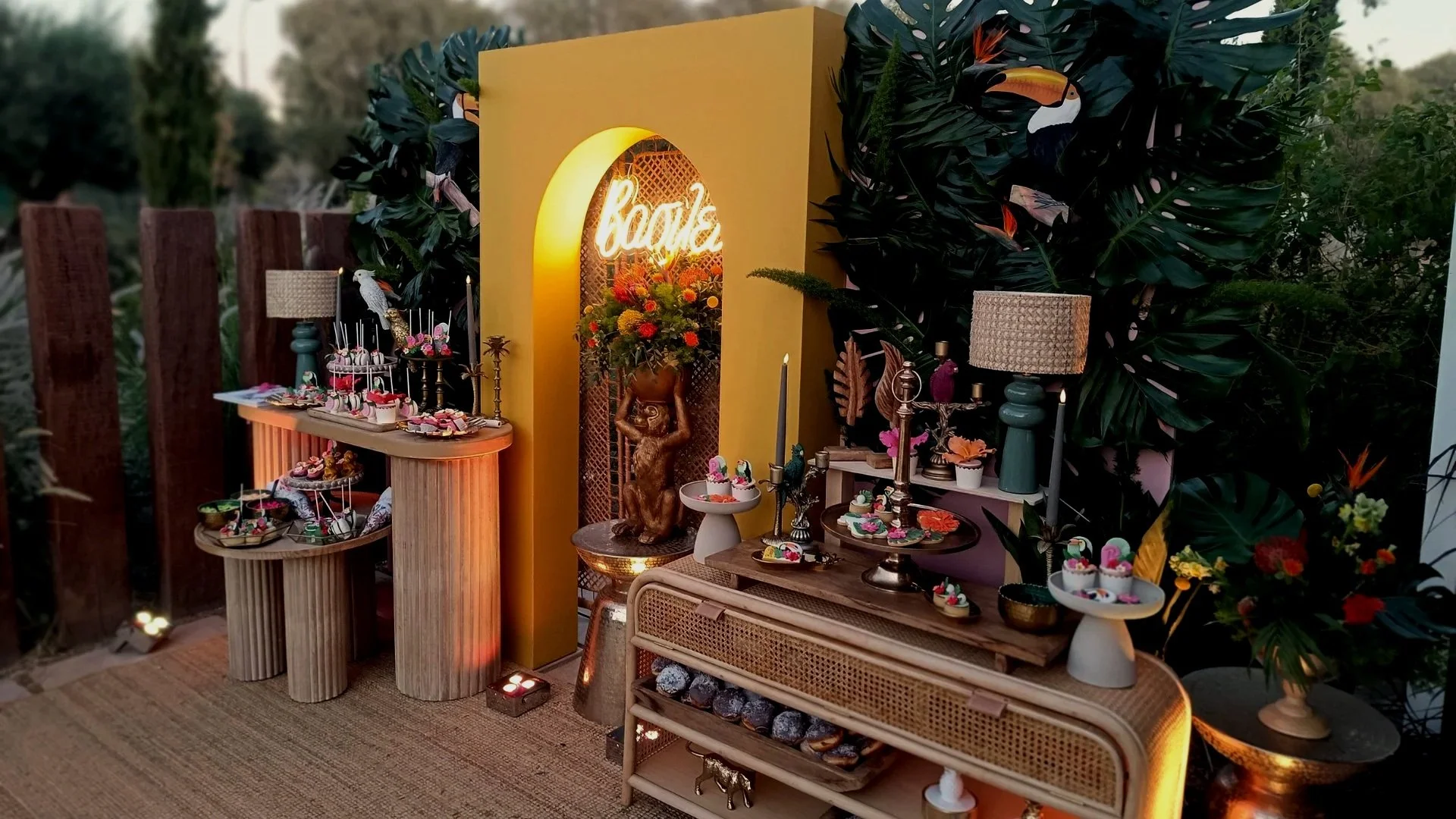 Decorative outdoor party setup featuring a yellow backdrop with a "bride" neon sign, tropical plants, floral arrangements, candles, and a variety of desserts displayed on tables.