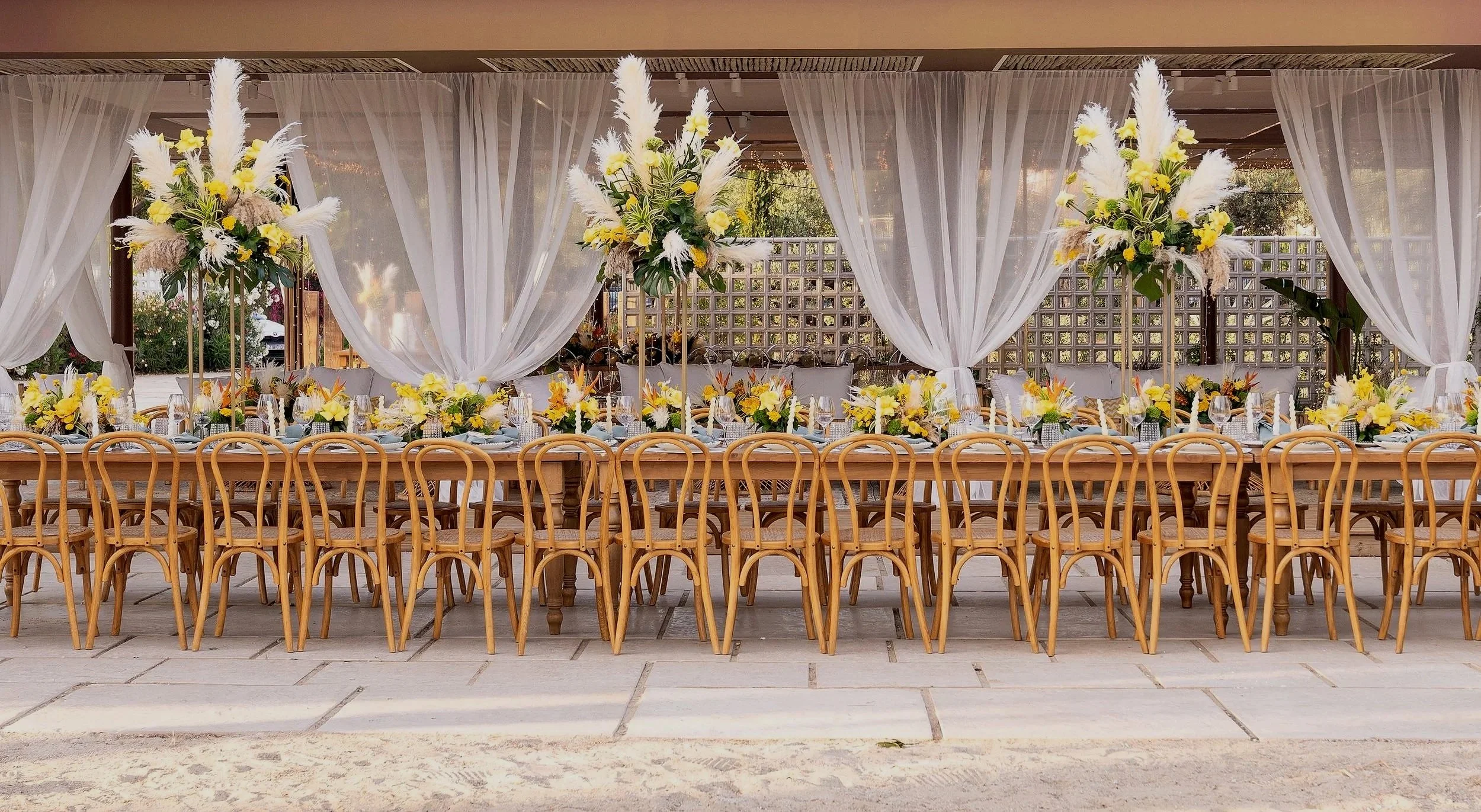 An elegantly decorated banquet table set for a celebration, featuring white curtains, floral centerpieces with yellow and white flowers, and wooden chairs surrounding the table.