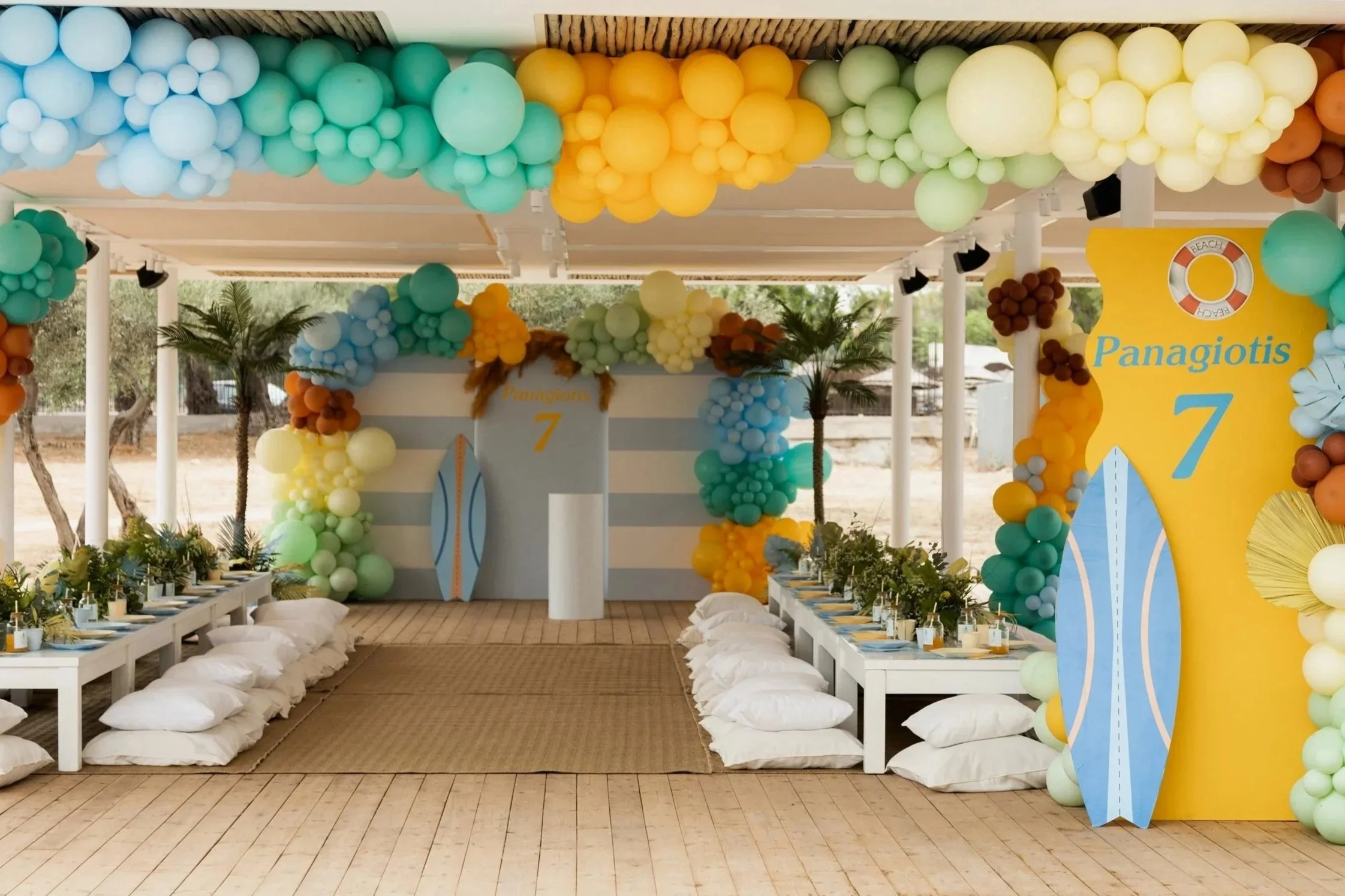 Beach-themed party setup with colorful balloon decorations, palm trees, and surfboard props. Long tables with pillows on the floor for seating and tropical centerpieces. Bright yellow and blue surfboard design with the text "Panagiotis 7" and a lifeb