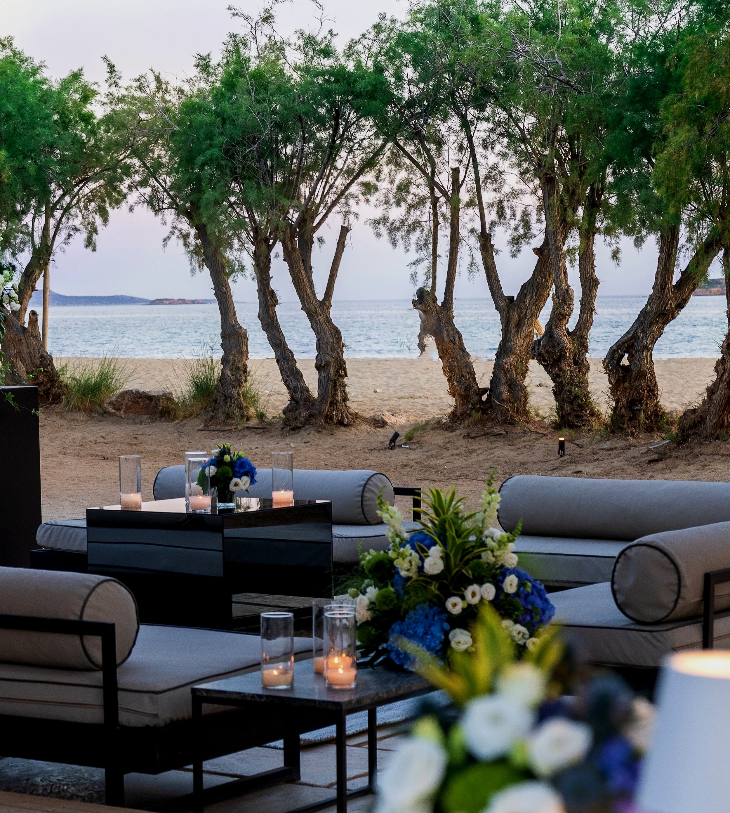 A beachside outdoor seating area with sofas and coffee tables decorated with candles and floral arrangements, overlooking the beach and water through a line of trees.