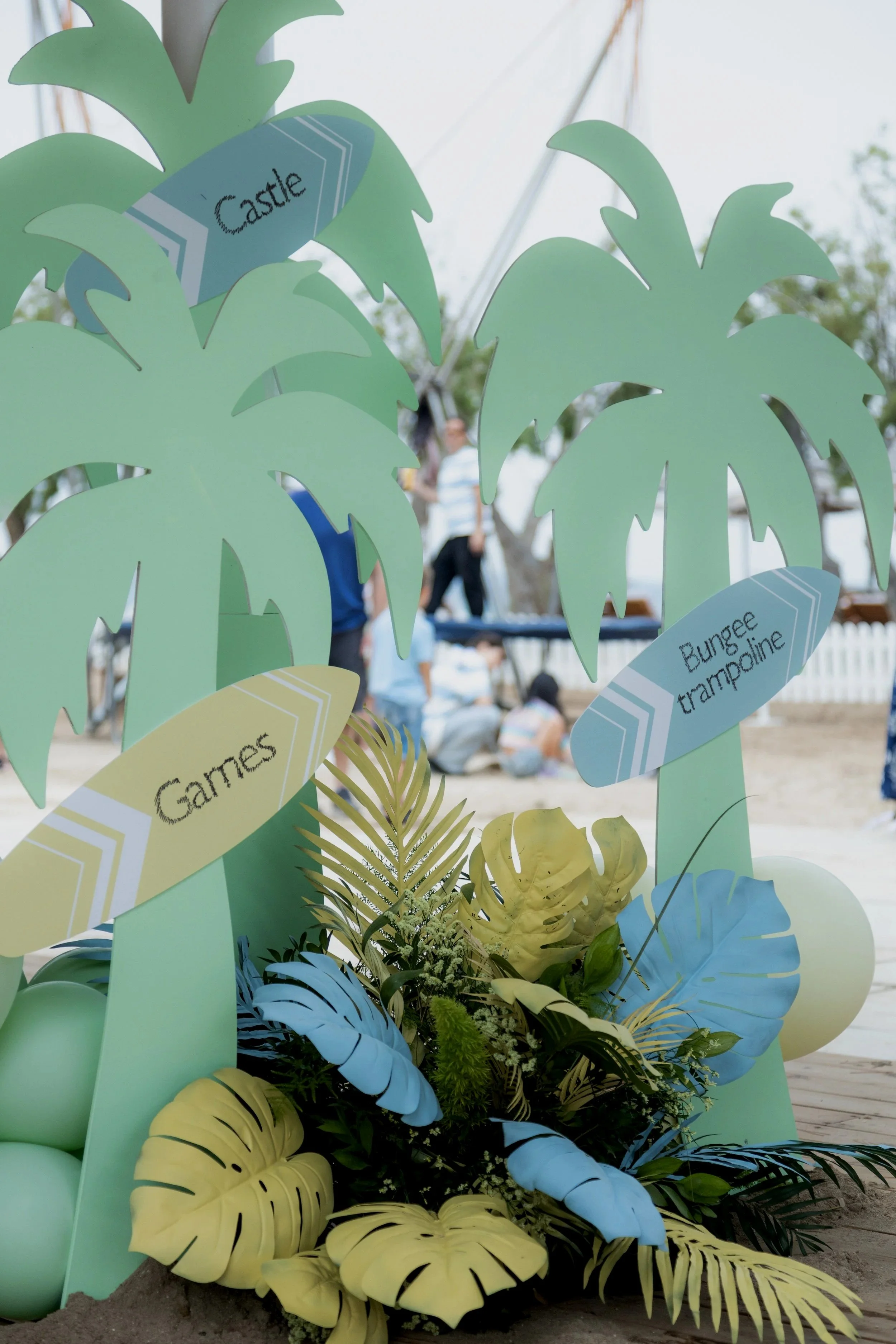 Decorative tropical theme display with paper palm trees labeled 'Castle,' 'Gates,' and 'Bungee Trampoline' at an outdoor event, with people and a tent in the background.
