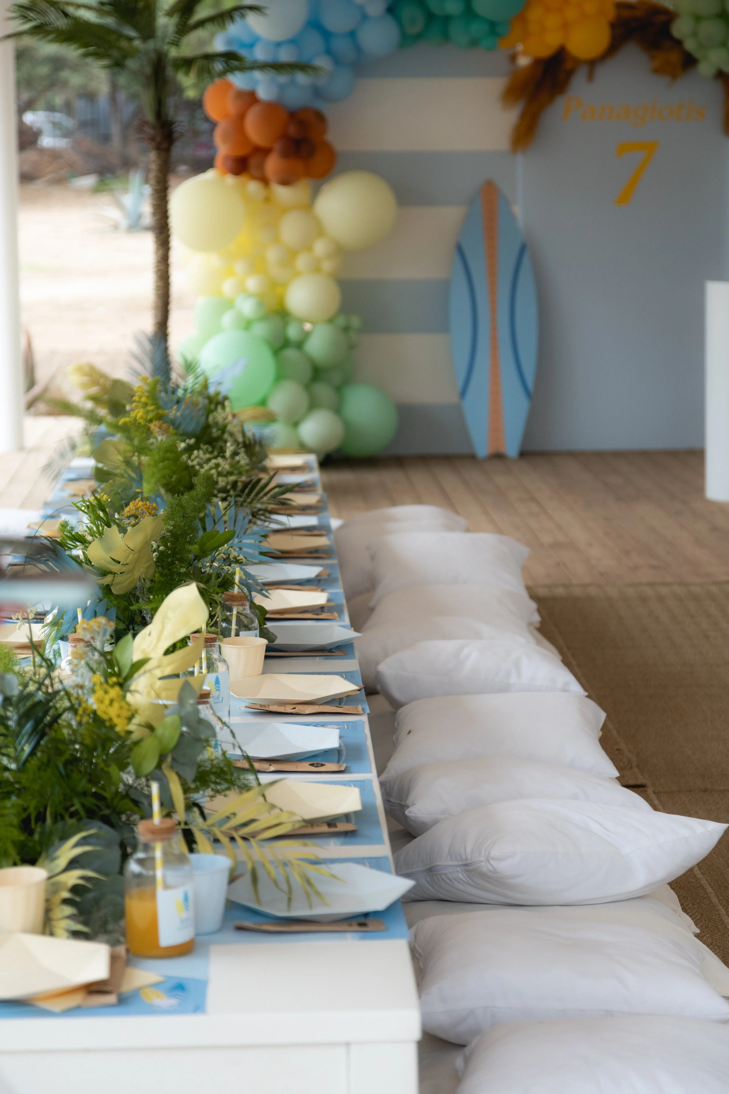 A decorated table set for a celebration with white pillows on the ground, floral arrangements, and a backdrop with balloons and a surfboard, indicating a beach or pool party themed event.