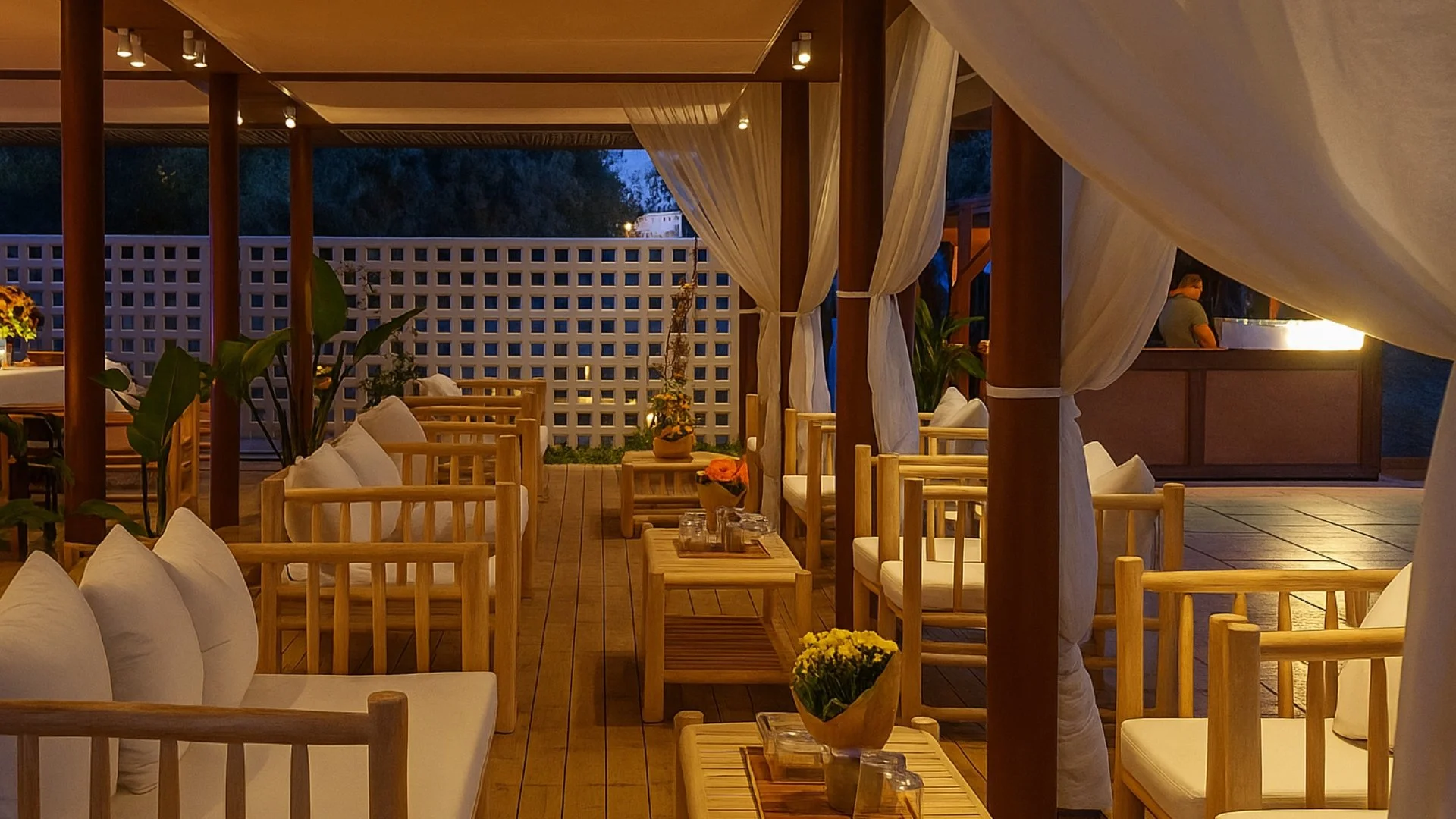 Empty restaurant patio with wooden chairs and tables, potted plants, soft lighting, and a counter at night