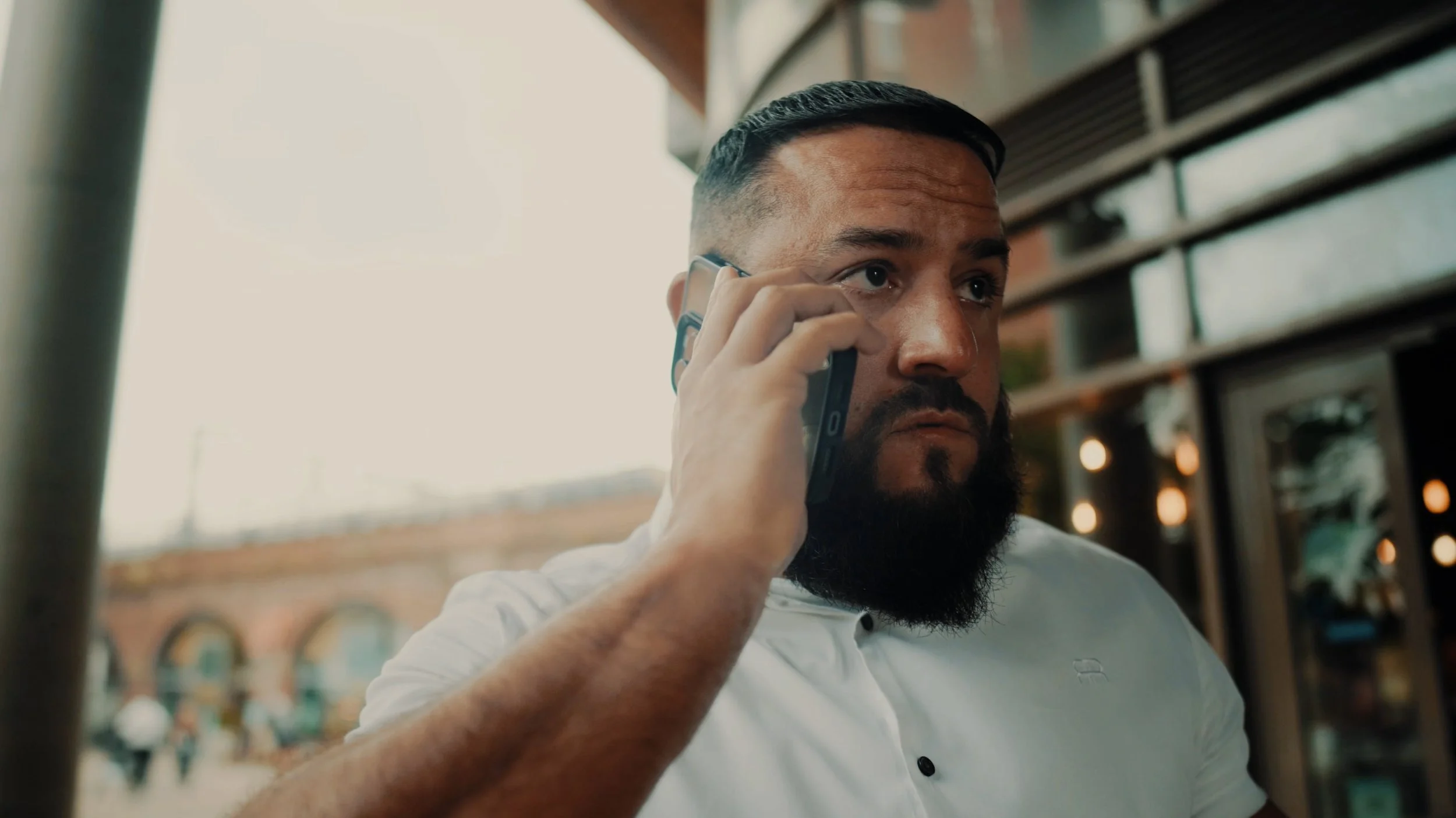 A man with a beard and short hair talking on a mobile phone outside a building