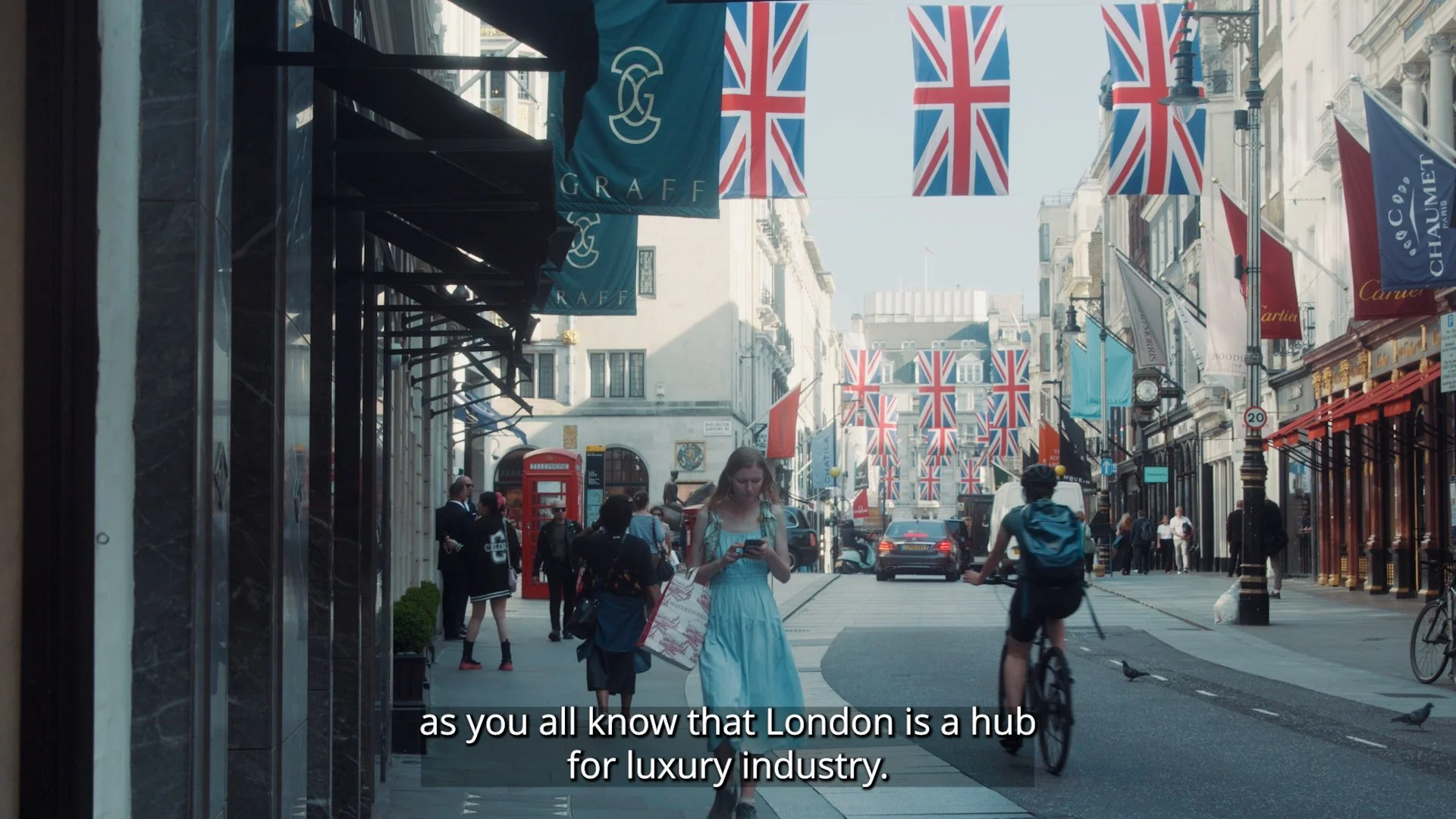A busy London street decorated with Union Jack flags hanging overhead. People walking, shopping, and riding bikes, with luxury store signs visible.