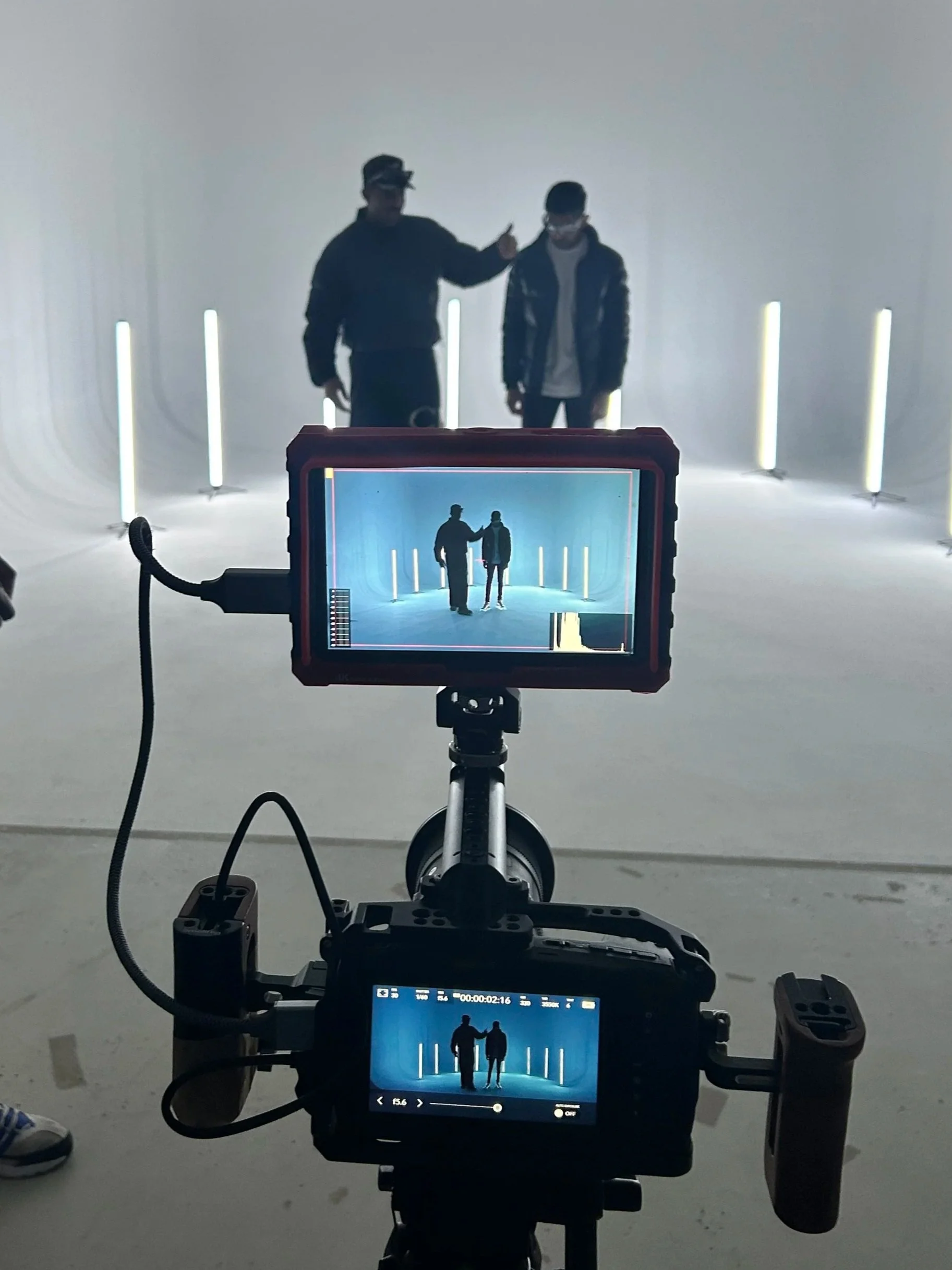 Two people standing in front of a white studio backdrop with vertical light tubes. A camera on a tripod is filming them, and its screen shows their silhouette against the light tubes.