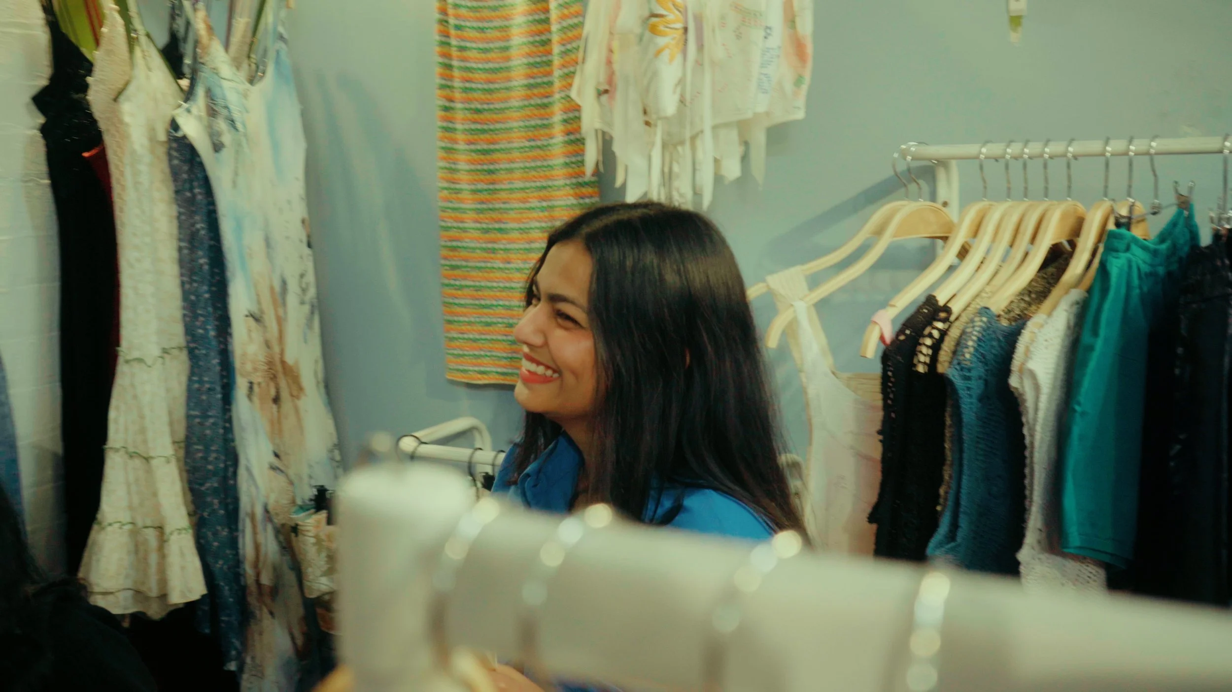 A woman with long dark hair smiling in a clothing store, surrounded by hanging dresses and tops on racks.
