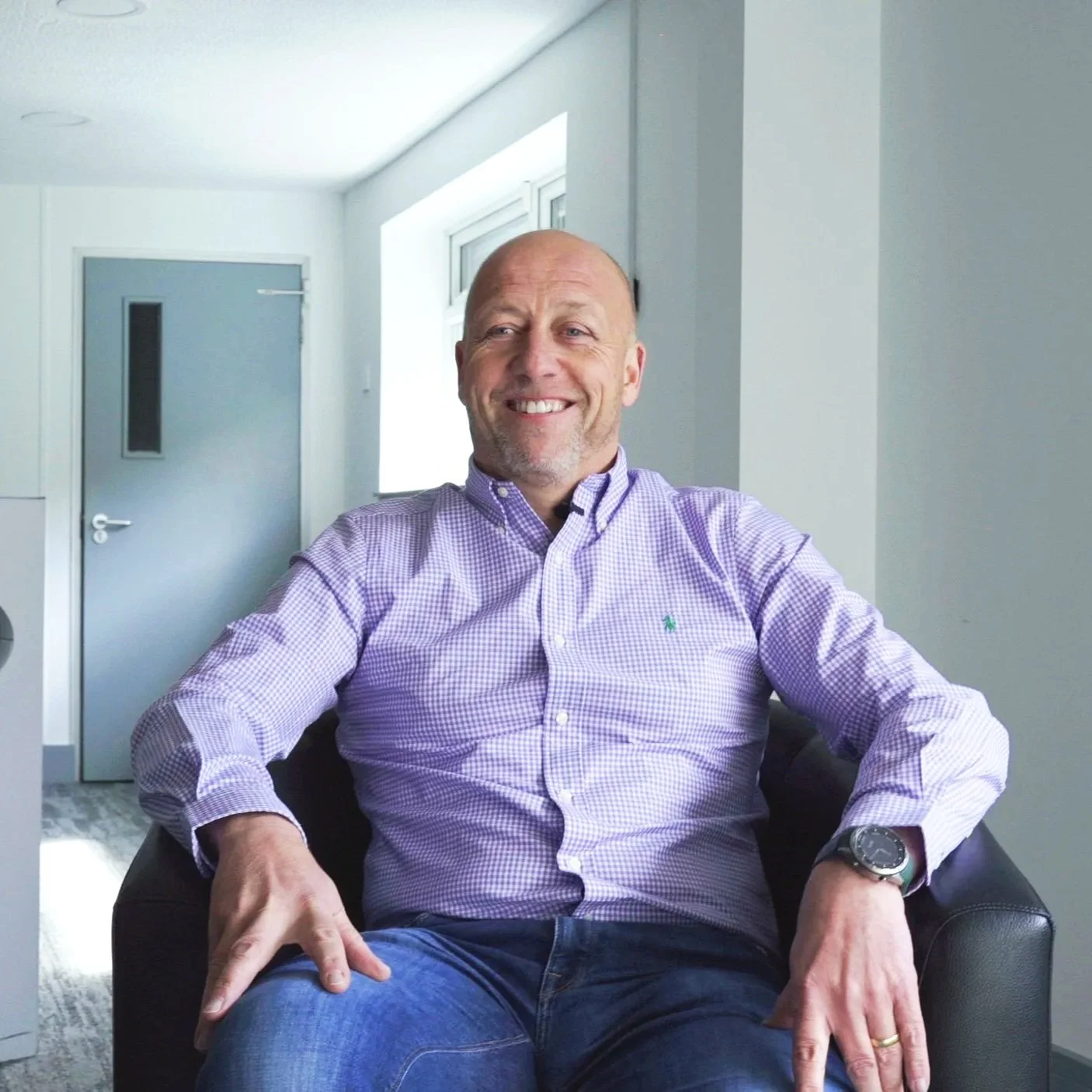 A middle-aged man with a bald head and a beard, smiling, is sitting on a black leather chair indoors. He's wearing a light purple checkered shirt and a wristwatch, with his hands resting on his legs.