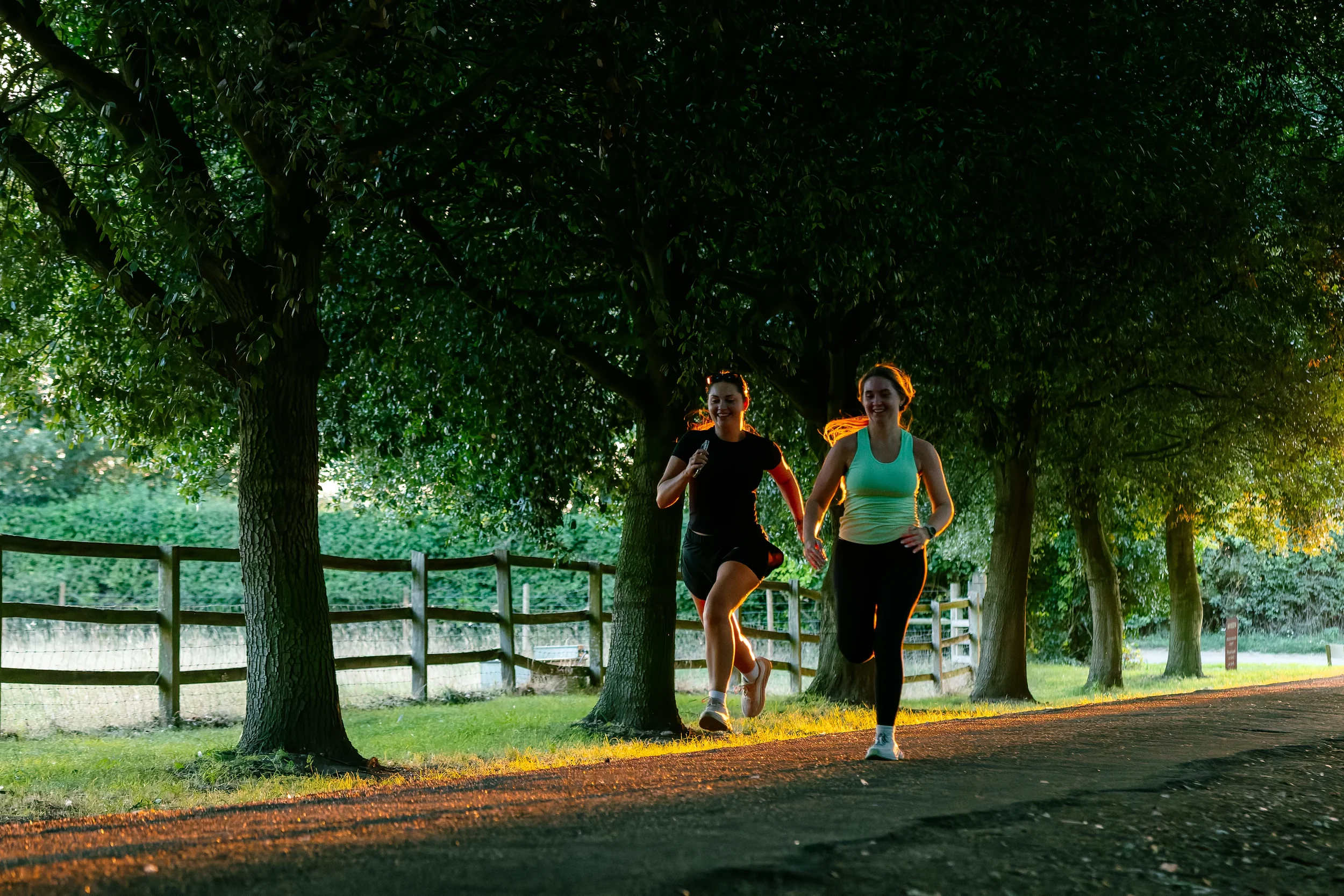 Two women jogging on a tree-lined path during sunset, smiling and wearing athletic clothes.
