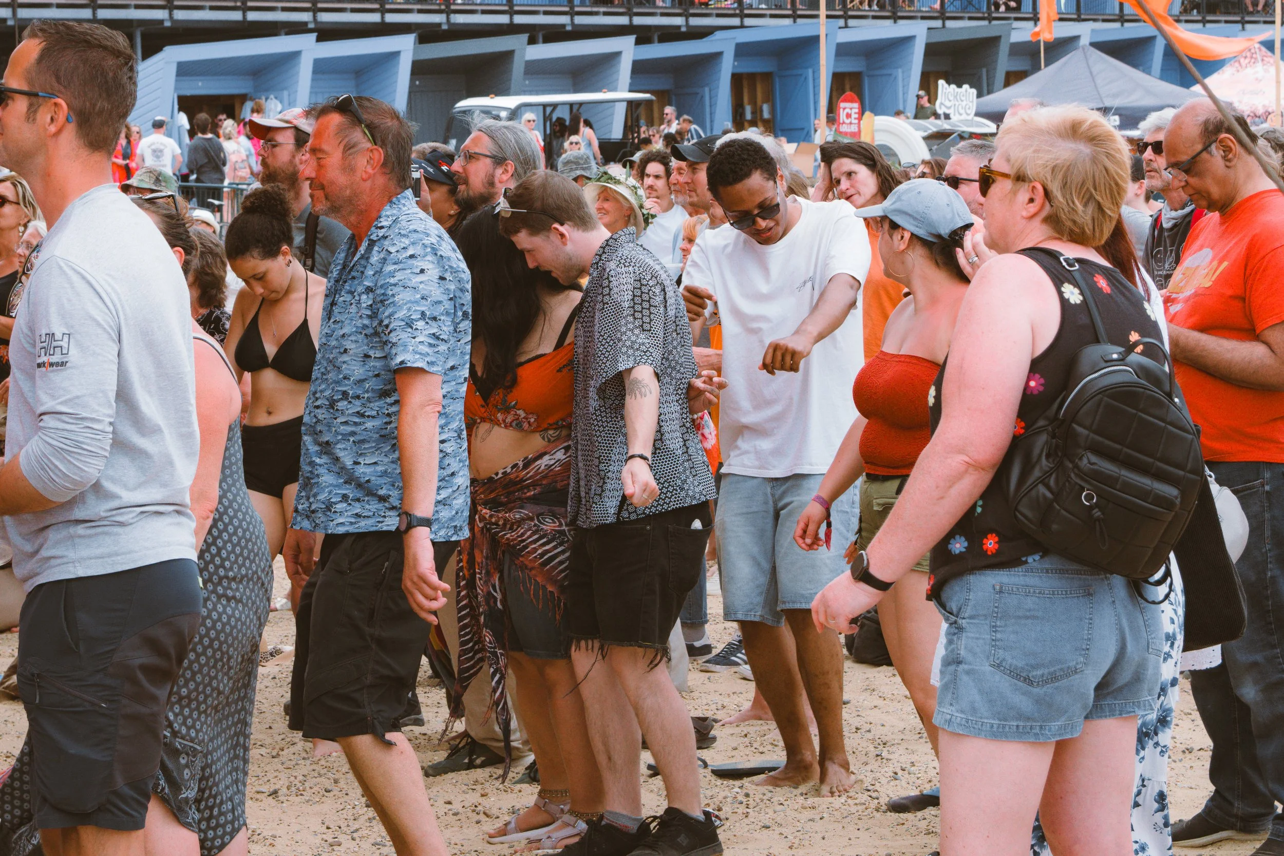 People standing in line outdoors on a sandy surface at a festival or event, some wearing summer clothing, in front of a blue structure with tents and signage.