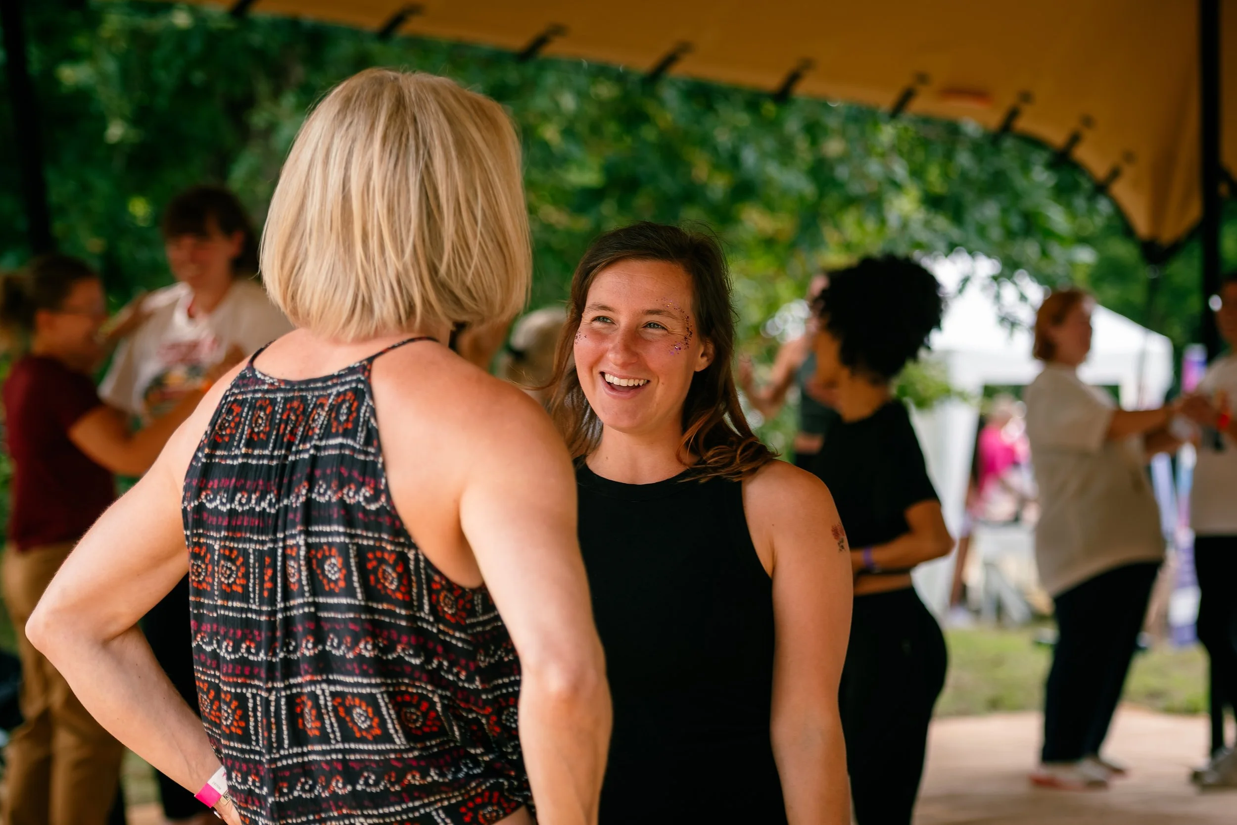 Two women talking and smiling at an outdoor event under a canopy, with other people socializing in the background.