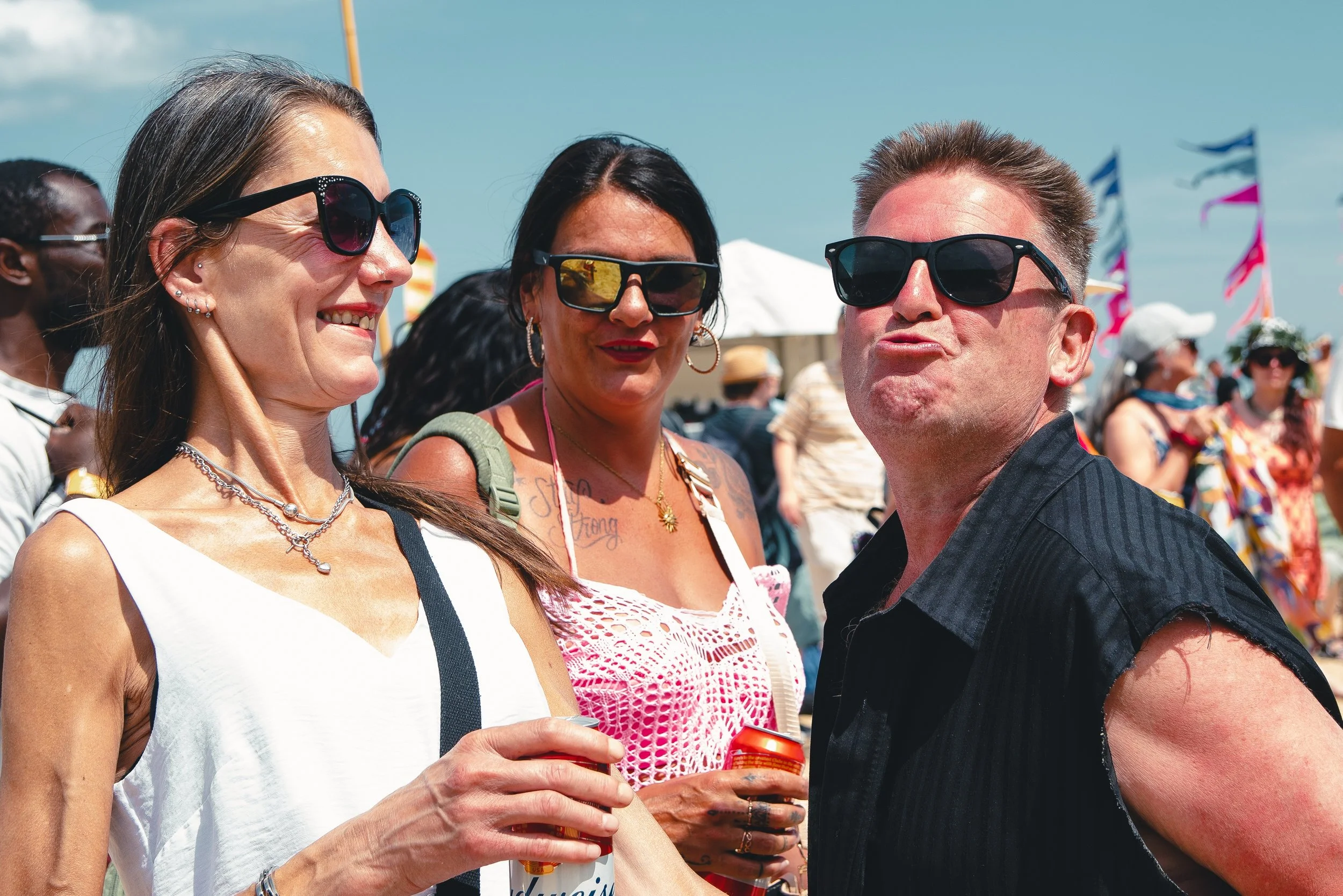 Three people at an outdoor event, two women and one man, wearing sunglasses and casual summer clothing, with a crowd, tents, and flags in the background.