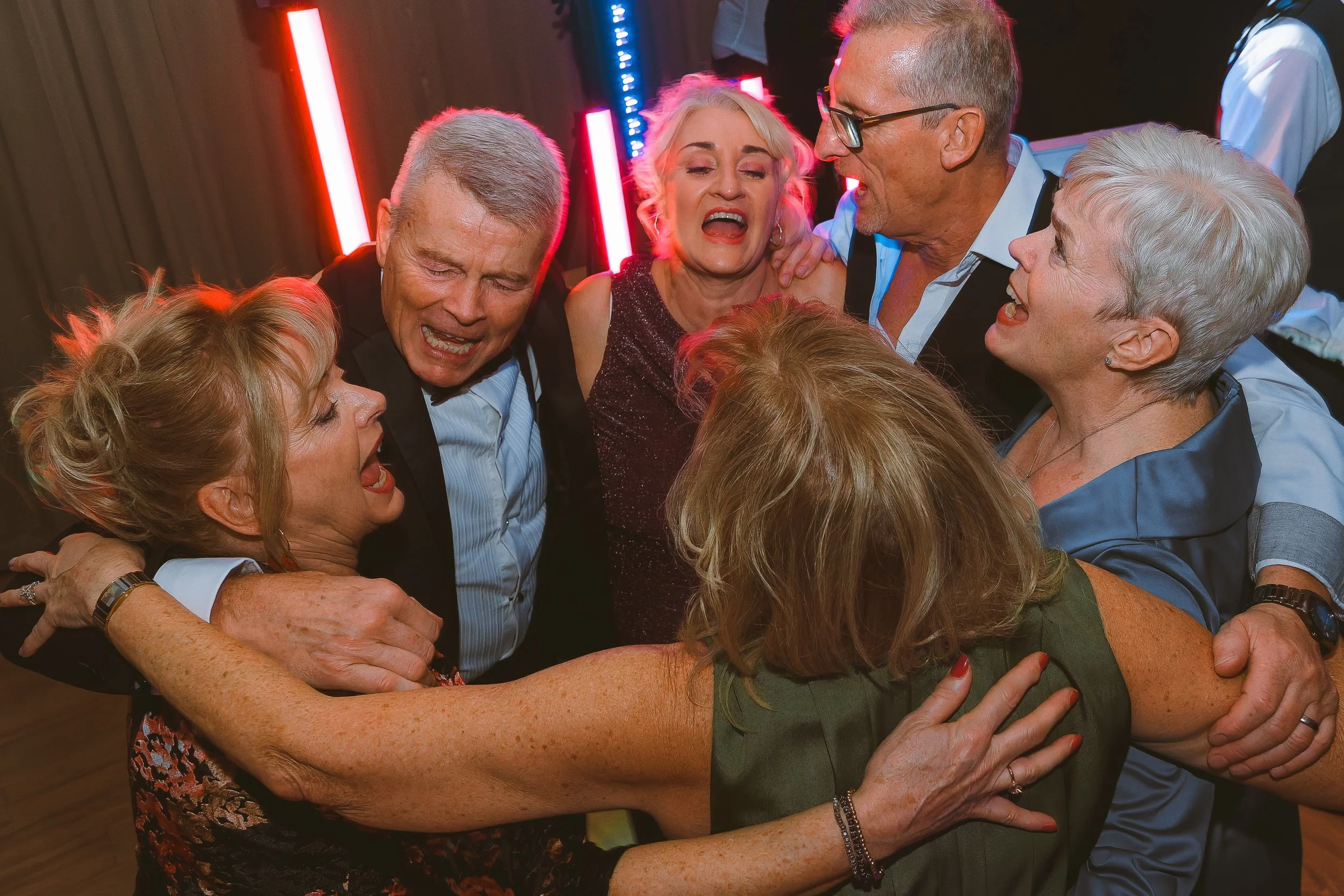 Group of elderly people hugging and celebrating together at a party or gathering, with bright lighting and a festive atmosphere.