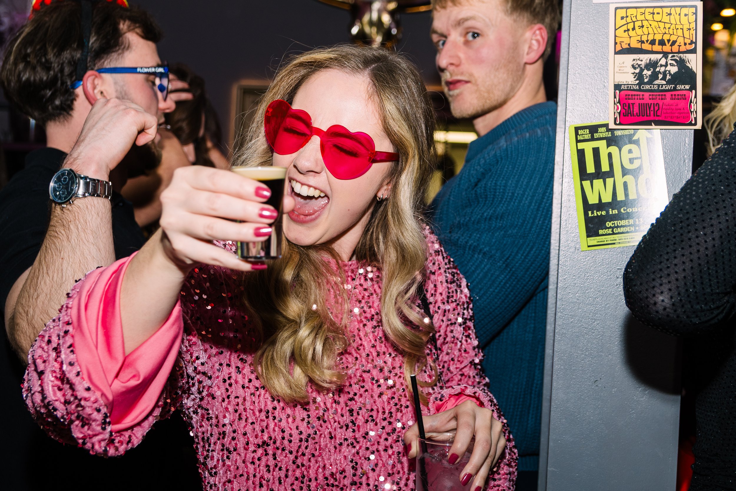 A woman with long wavy hair wearing party glasses and a pink sequined top smiling and holding a shot glass at a party.