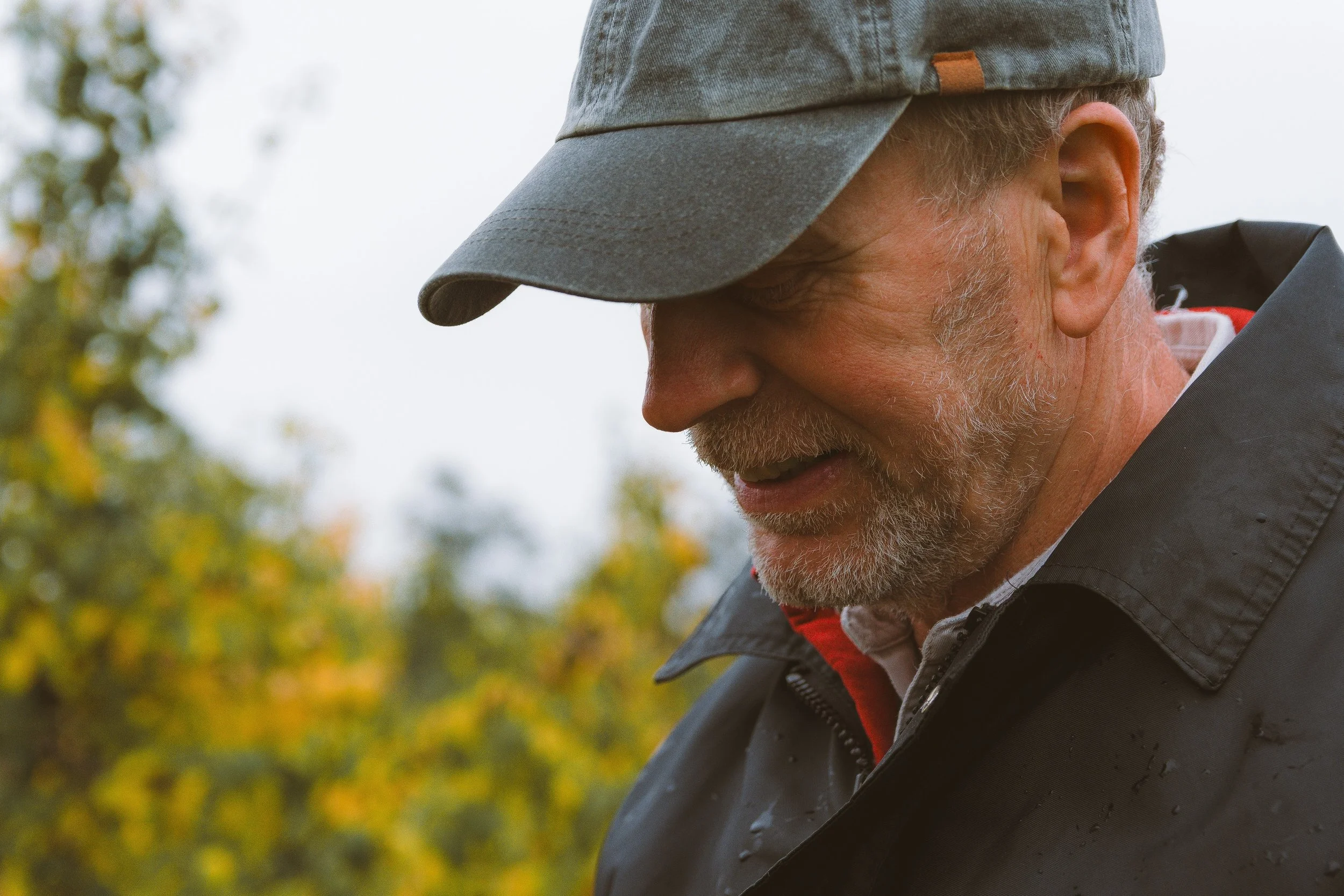 Close-up of a smiling older man wearing a gray cap and black jacket outdoors with autumn foliage in the background.