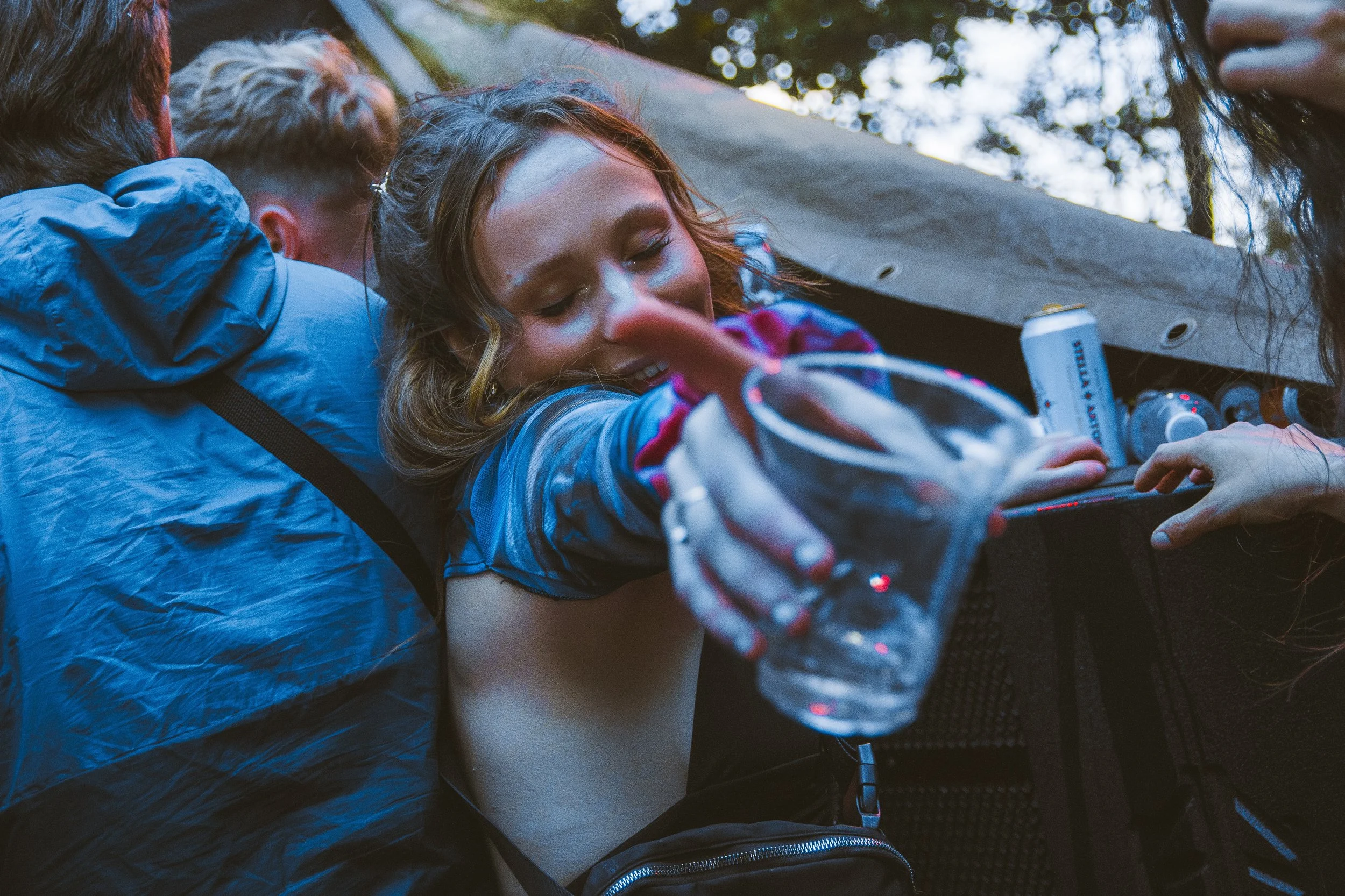 A young woman reaching out with a clear plastic water bottle, surrounded by people outdoors, with a tent or canopy overhead and trees in the background.