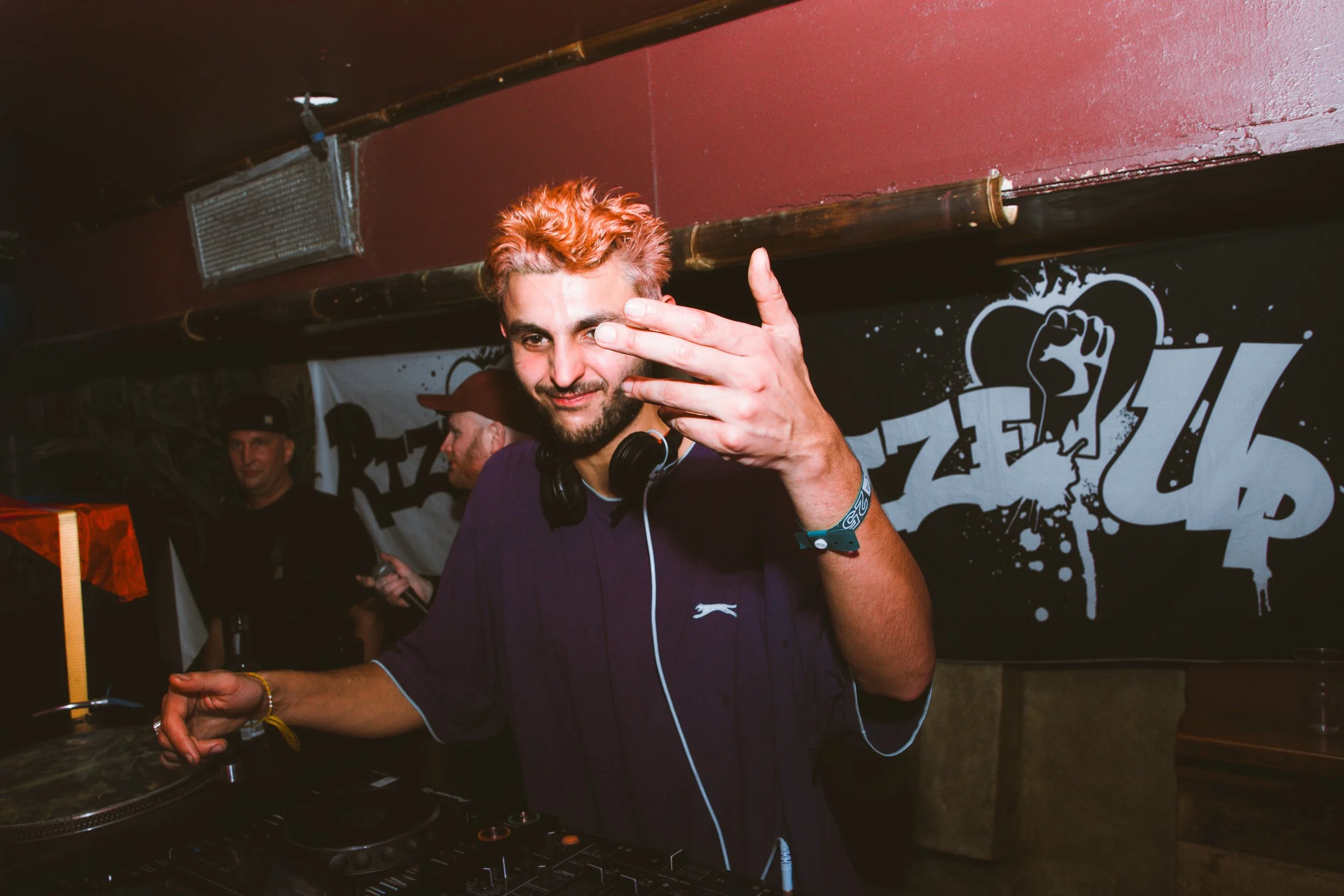 A DJ with reddish hair and a beard, wearing a dark shirt, standing behind a DJ mixer with headphones around his neck, gesturing toward the camera with his right hand, in a club with graffiti-style wall art in the background.