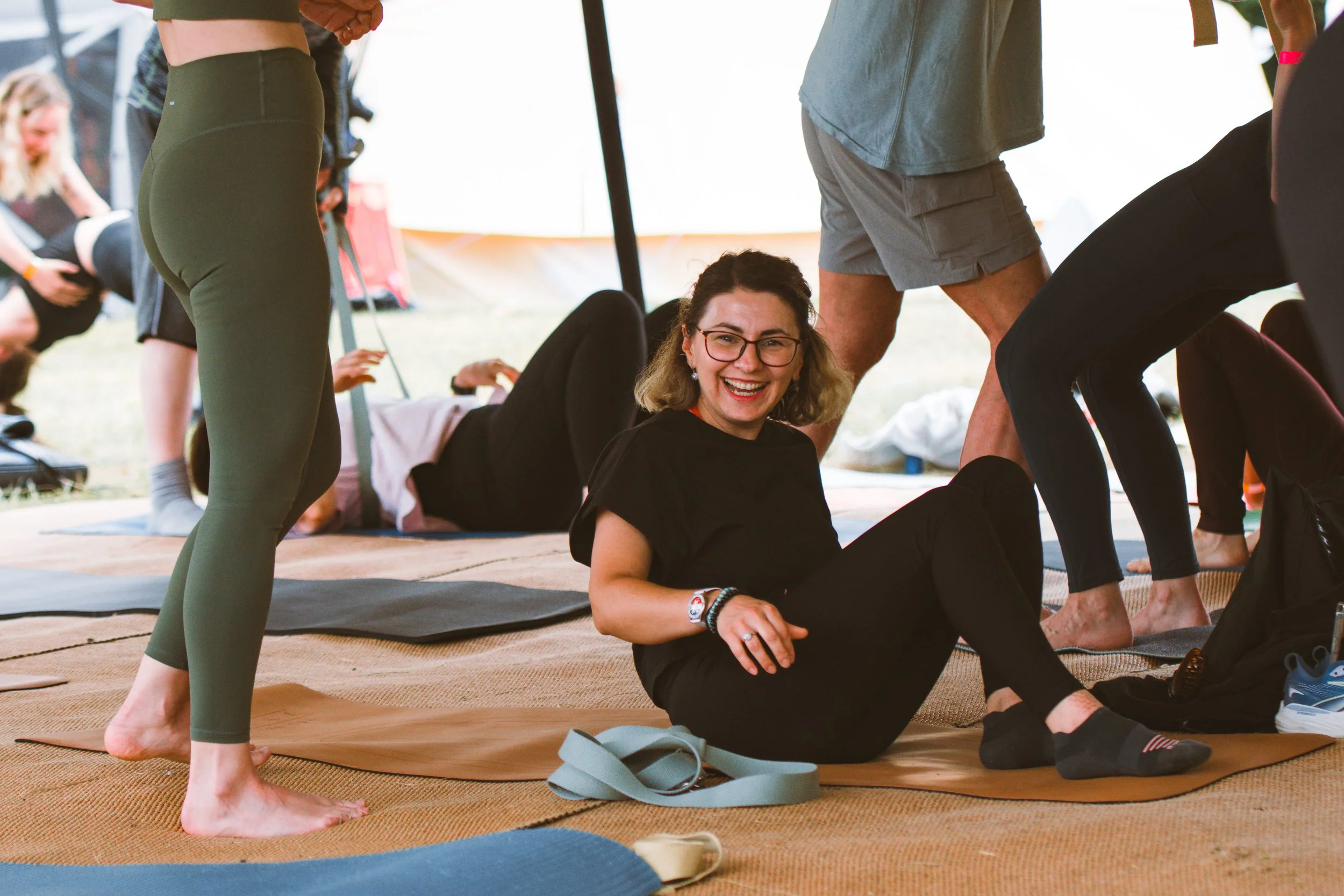 A woman with glasses and a black shirt sitting on a yoga mat in a group yoga class, smiling at the camera. People around her are in various yoga poses or preparing for yoga practice.