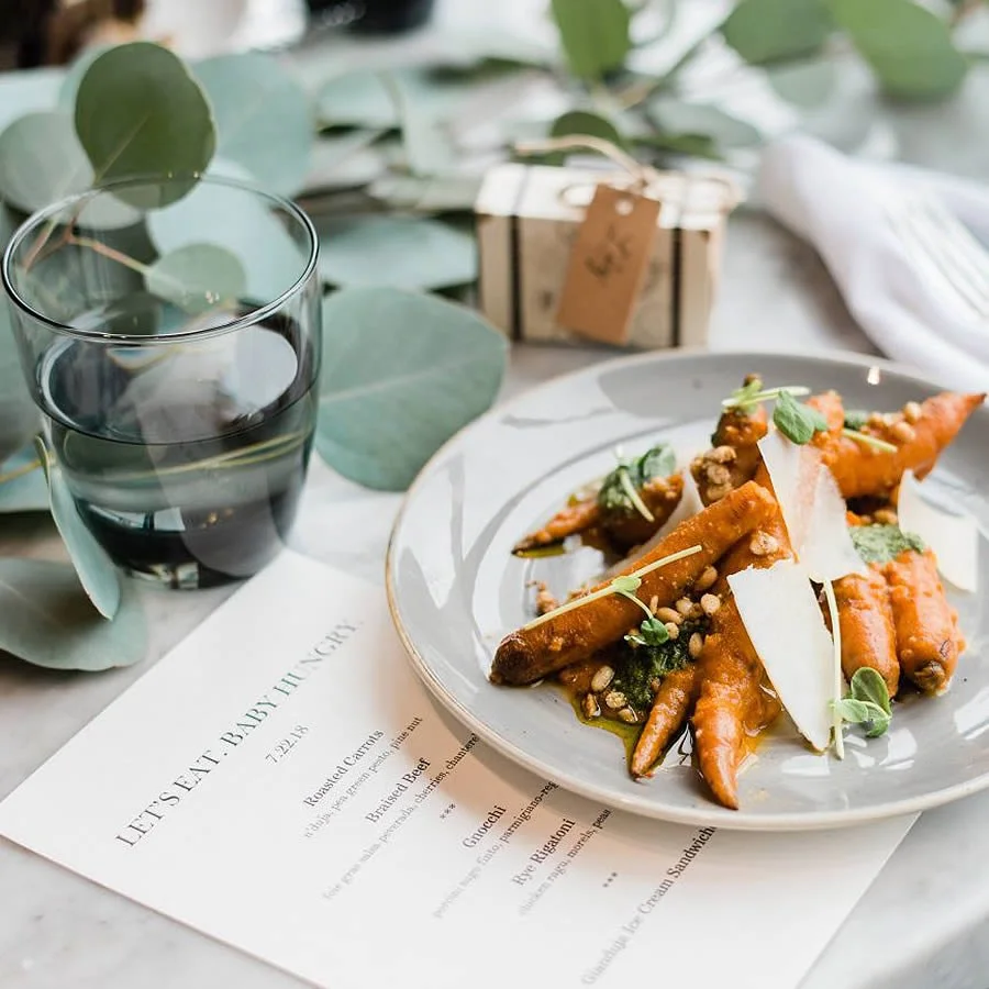 Plate of roasted carrots garnished with microgreens and cheese shavings, two glasses of water, a gift box, and a menu with the heading