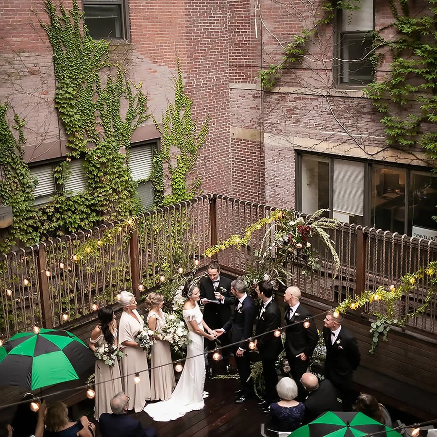 Outdoor wedding ceremony with bride and groom exchanging vows, surrounded by bridesmaids and groomsmen, on a decorated wooden deck enclosed by a brick wall and string lights, with guests seated and standing, some holding umbrellas.