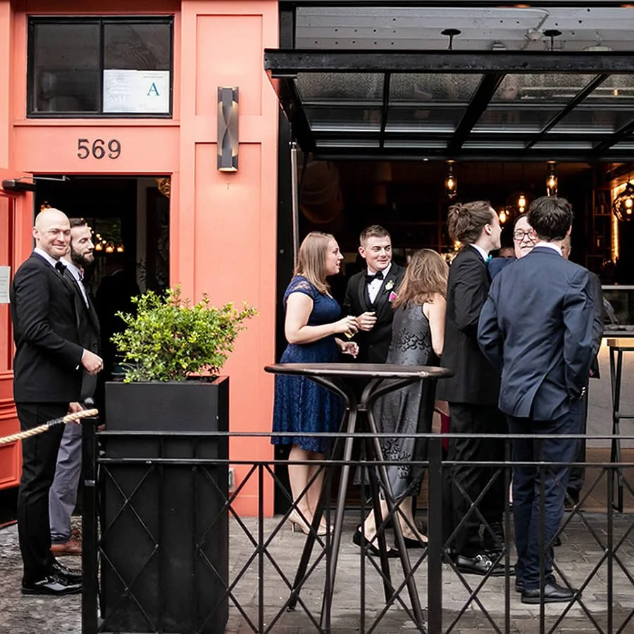 Group of people in formal attire chatting outside a restaurant with the address 569 on the wall.