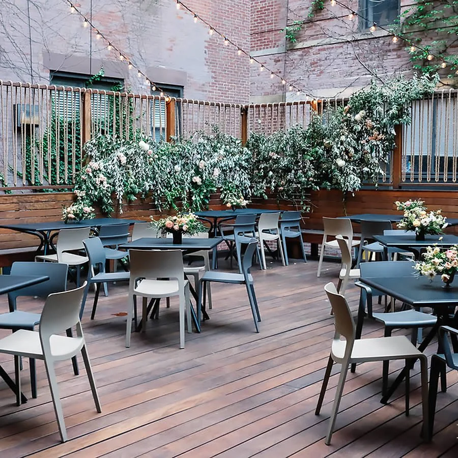 Outdoor patio with wooden deck, black and gray tables, and matching chairs. Decorative flowers in vases on tables. Greenery and flowering plants along the fence, with string lights overhead.