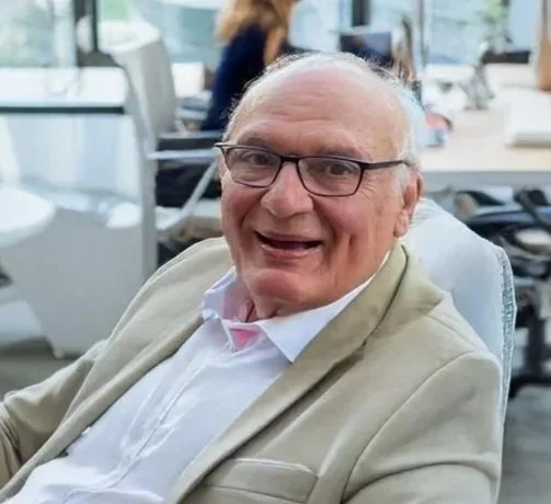 Smiling elderly man with glasses, wearing a beige blazer and white shirt, sitting at a desk in an office with large windows and other people working in the background.