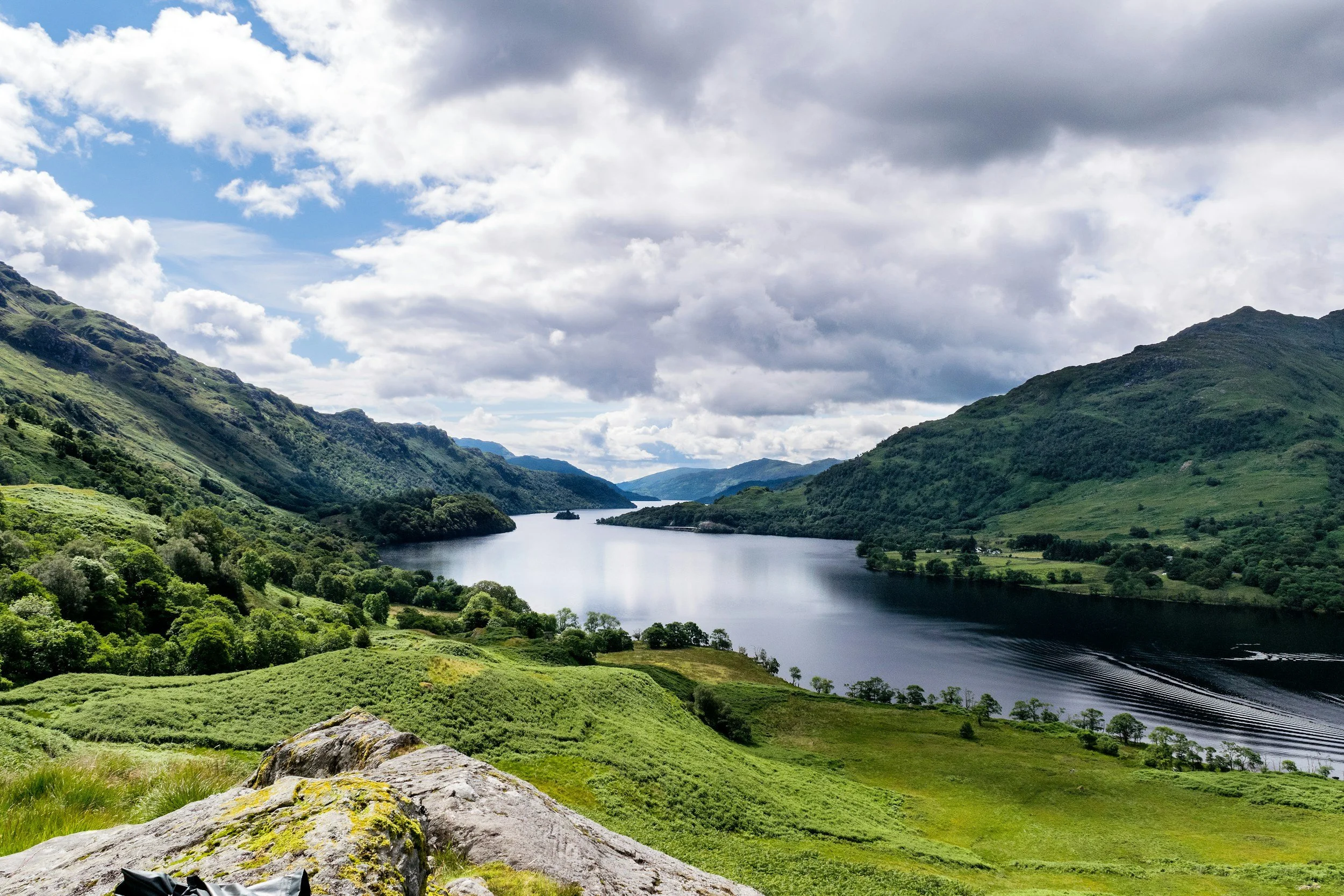 A scenic view of a lake surrounded by green hills and mountains under a partly cloudy sky.