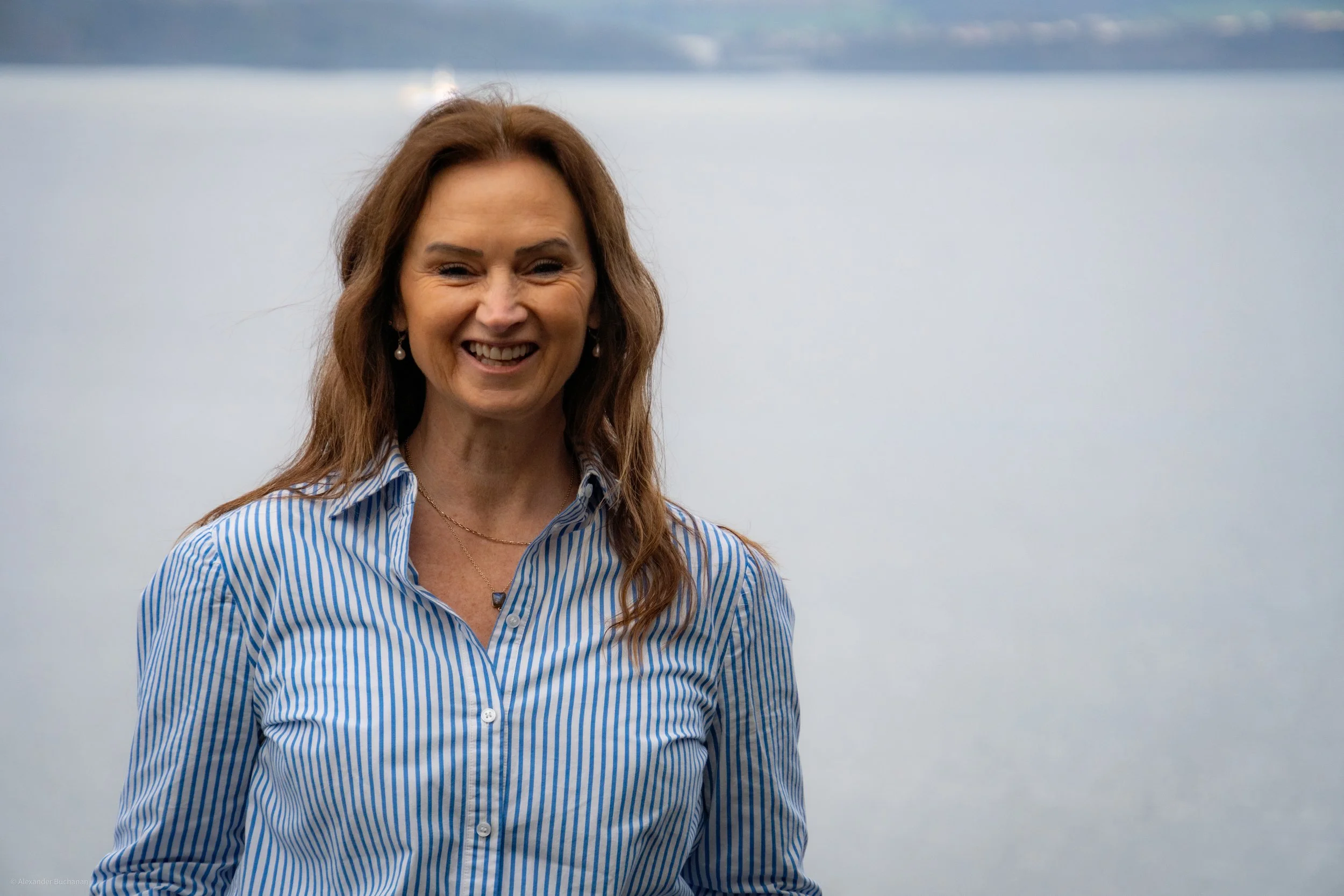 A woman (Joy Buchanan) with long brown hair smiling outdoors near a large body of water, wearing a blue and white striped shirt and jewelry.