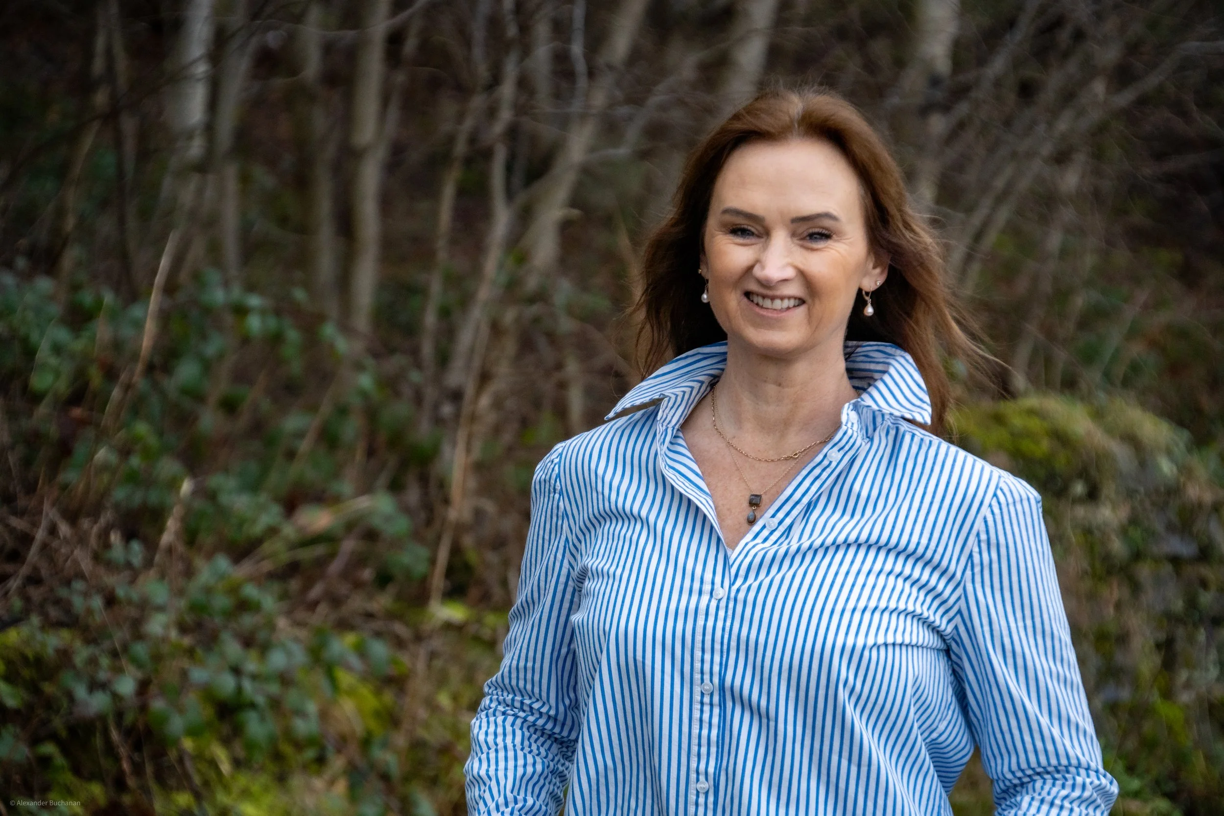 A woman (Joy Buchanan) with brown hair, smiling, wearing a blue and white striped shirt, earrings, and necklaces, standing outdoors in a natural setting with trees and greenery in the background.