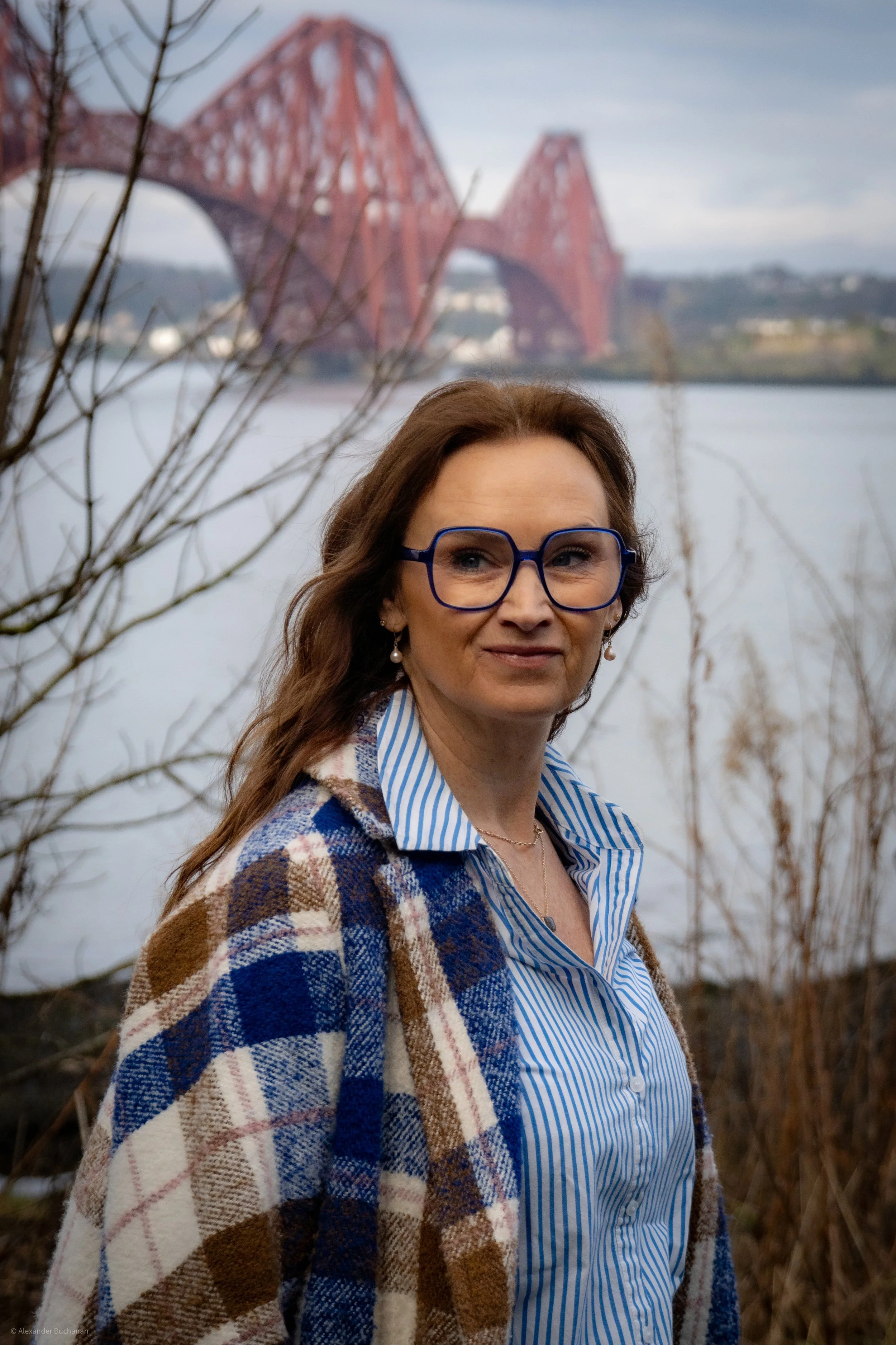 A woman (Joy Buchanan) with long brown hair, wearing glasses, a plaid brown and blue coat, and a blue and white striped shirt, standing outdoors with a body of water and a red bridge in the background.