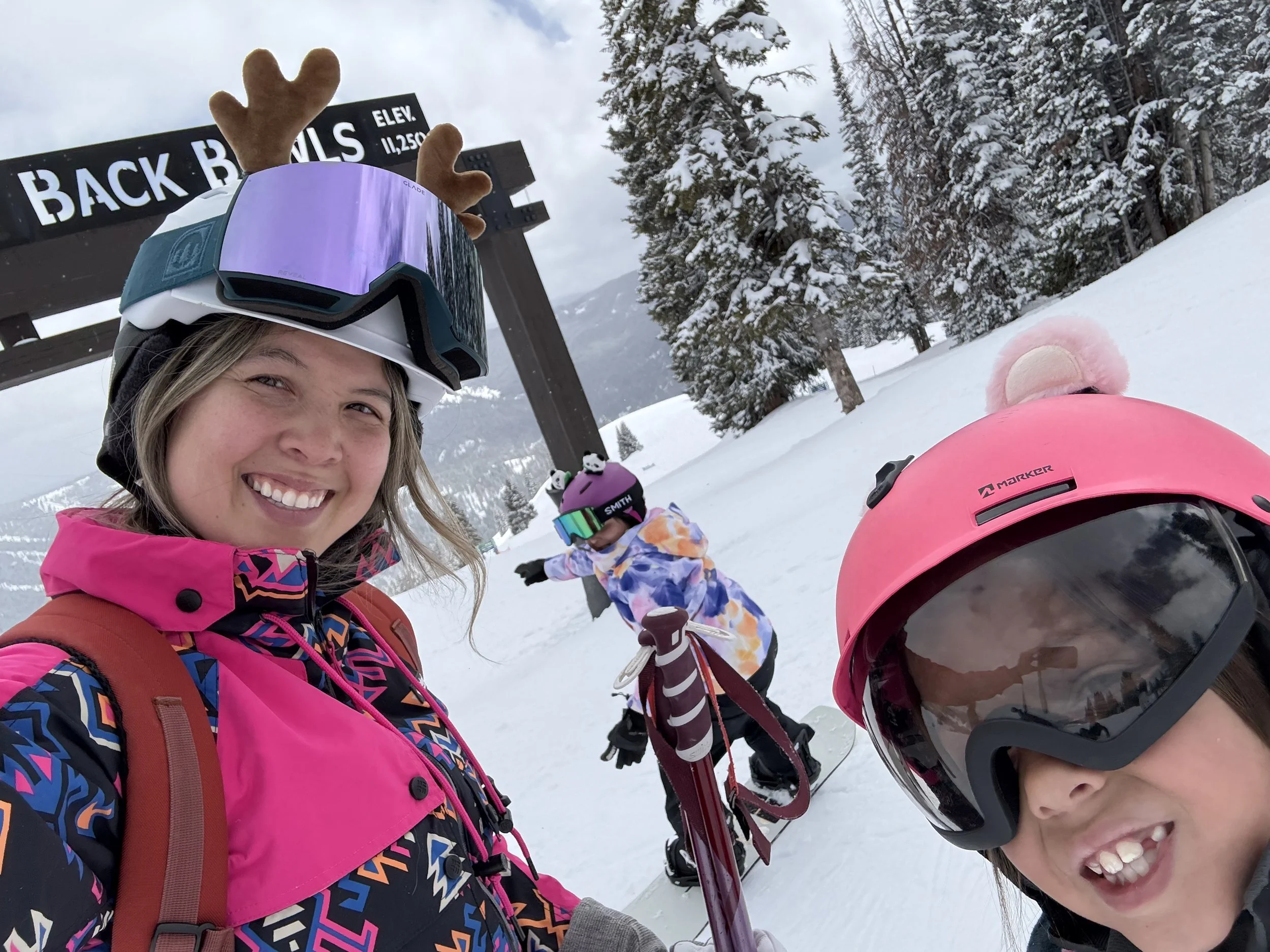 Smiling woman and children in colorful winter gear, skiing on a snow-covered mountain with trees and a sign that reads 'Back Bowls' in the background.